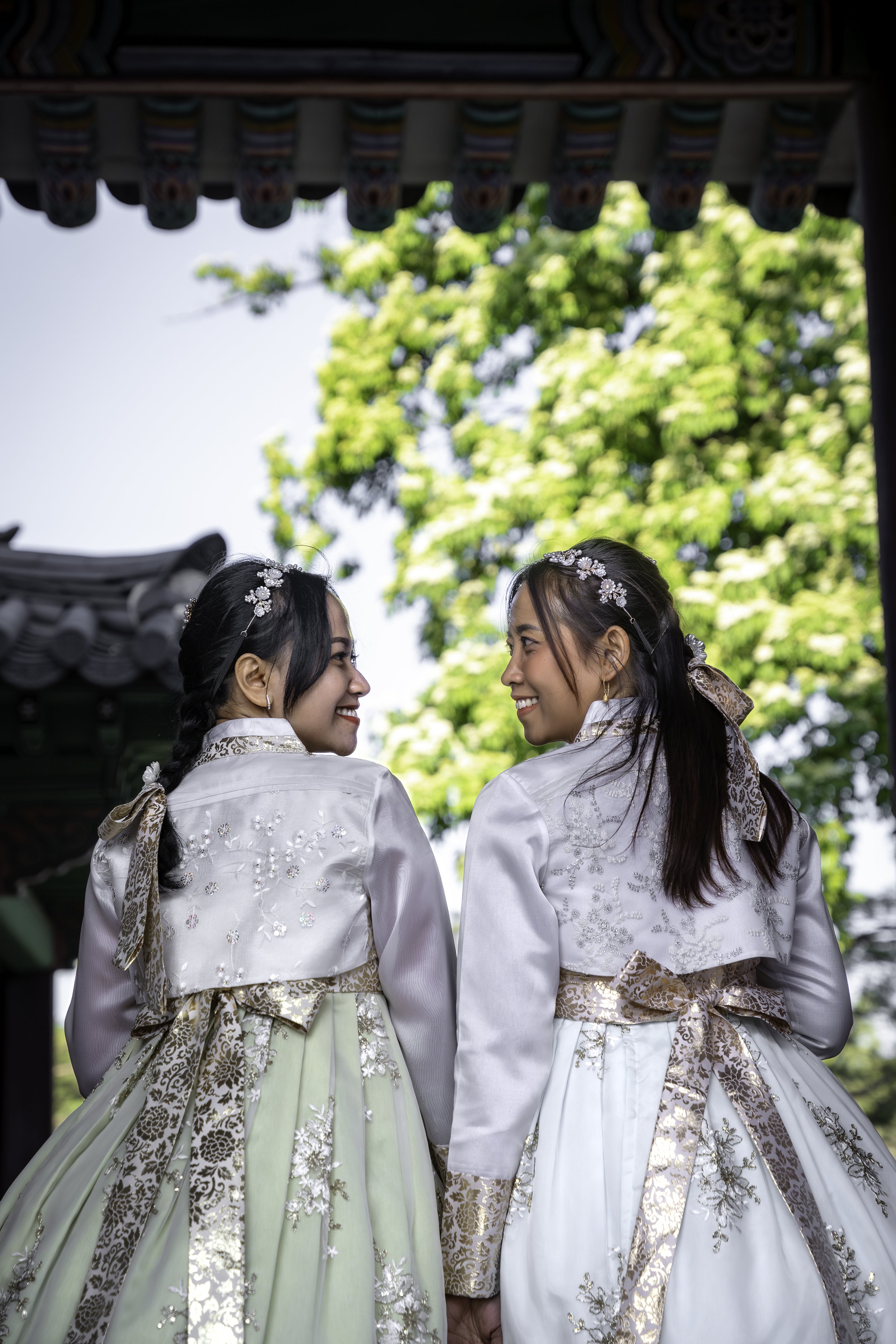 Two young women dressed in traditional Korean hanbok, smiling and looking at each other, outdoors with green trees and a building with Korean-style architecture in the background.