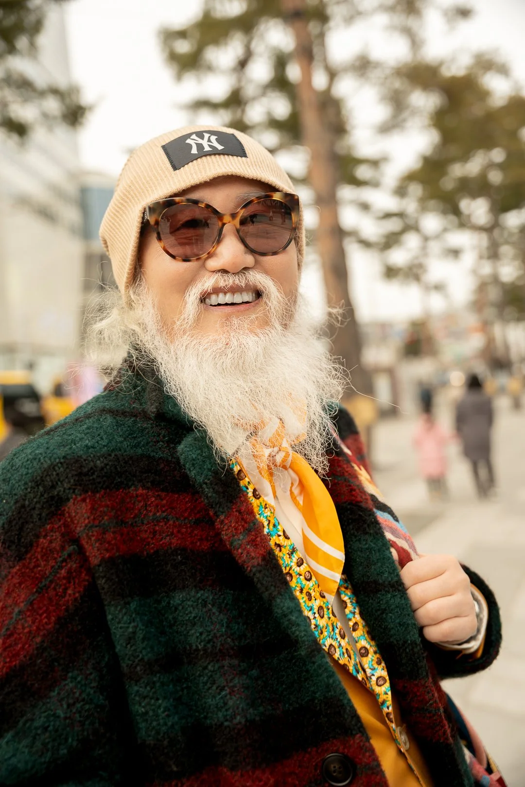 An elderly man with a long white beard and mustache, wearing large tortoiseshell sunglasses, a tan beanie with a NY logo, a colorful floral shirt, a yellow scarf, and a plaid jacket, smiling outdoors in a city street.