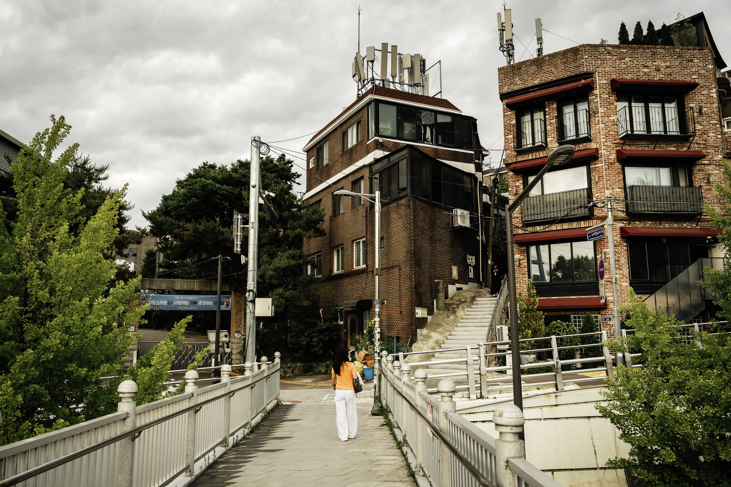 A woman walking on a pedestrian bridge with greenery on the sides, leading up to a cluster of brick apartment buildings with multiple antennas on the roof, under a cloudy sky.