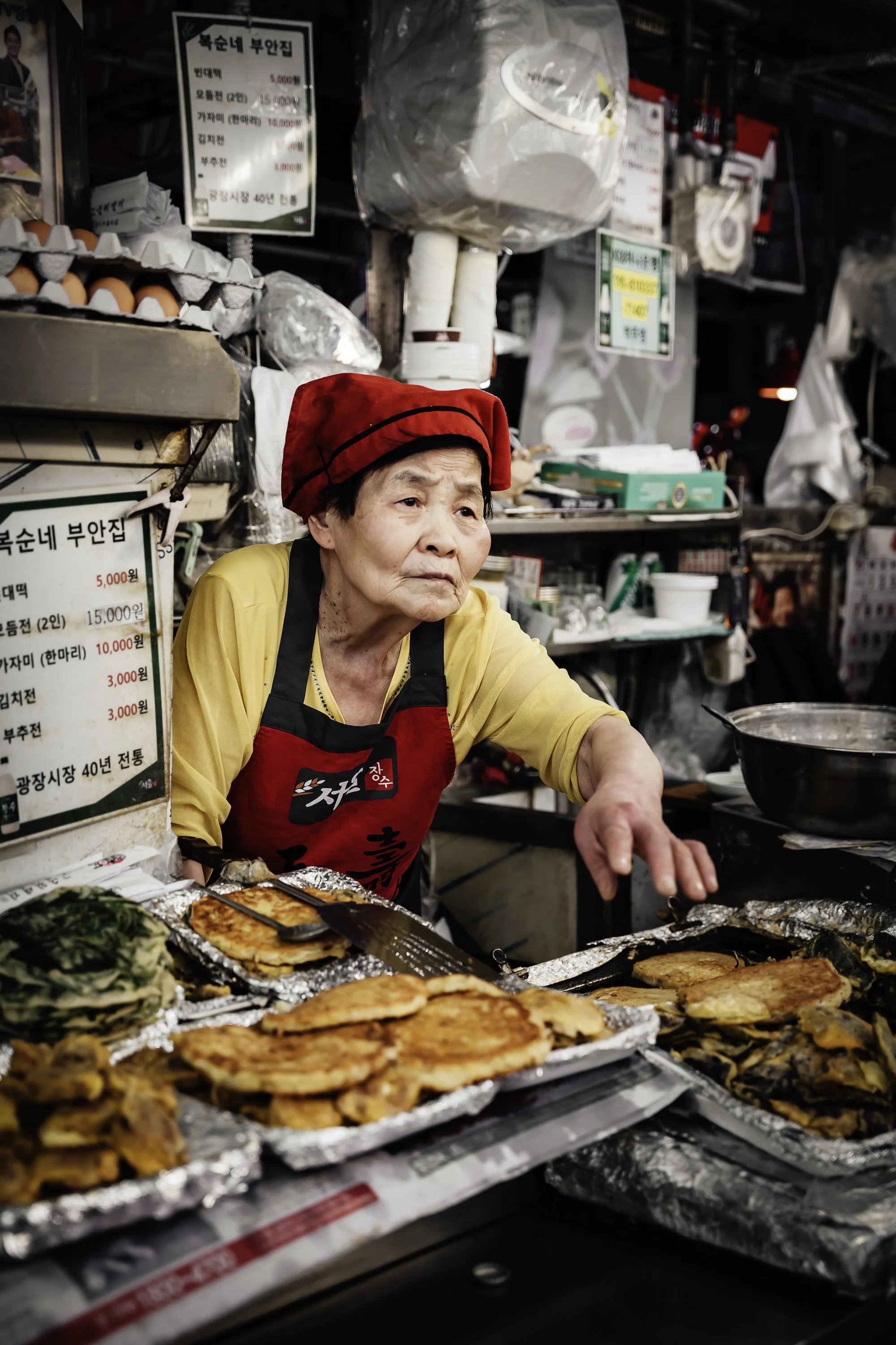 An elderly woman vendor wearing a red hat and apron working at a street food stall with various cooked dishes on display.
