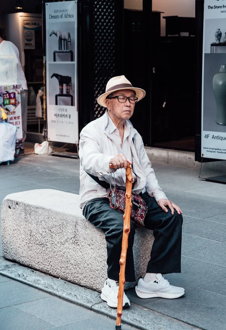 An elderly man with glasses, wearing a straw hat, white jacket, and black pants, sitting on a stone bench in an urban area. He is holding a walking stick and appears to be resting or sleeping with his eyes closed.