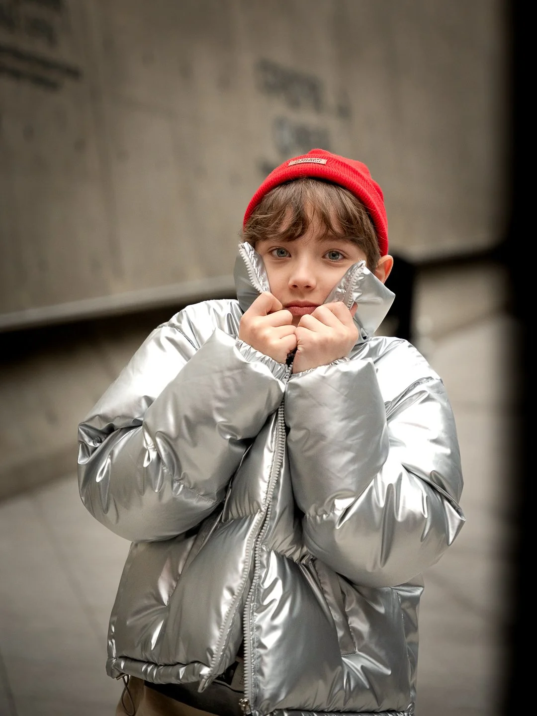 A young boy wearing a shiny silver puffer jacket and a red beanie hat, looking into the camera with a wistful expression, standing indoors against a neutral background.