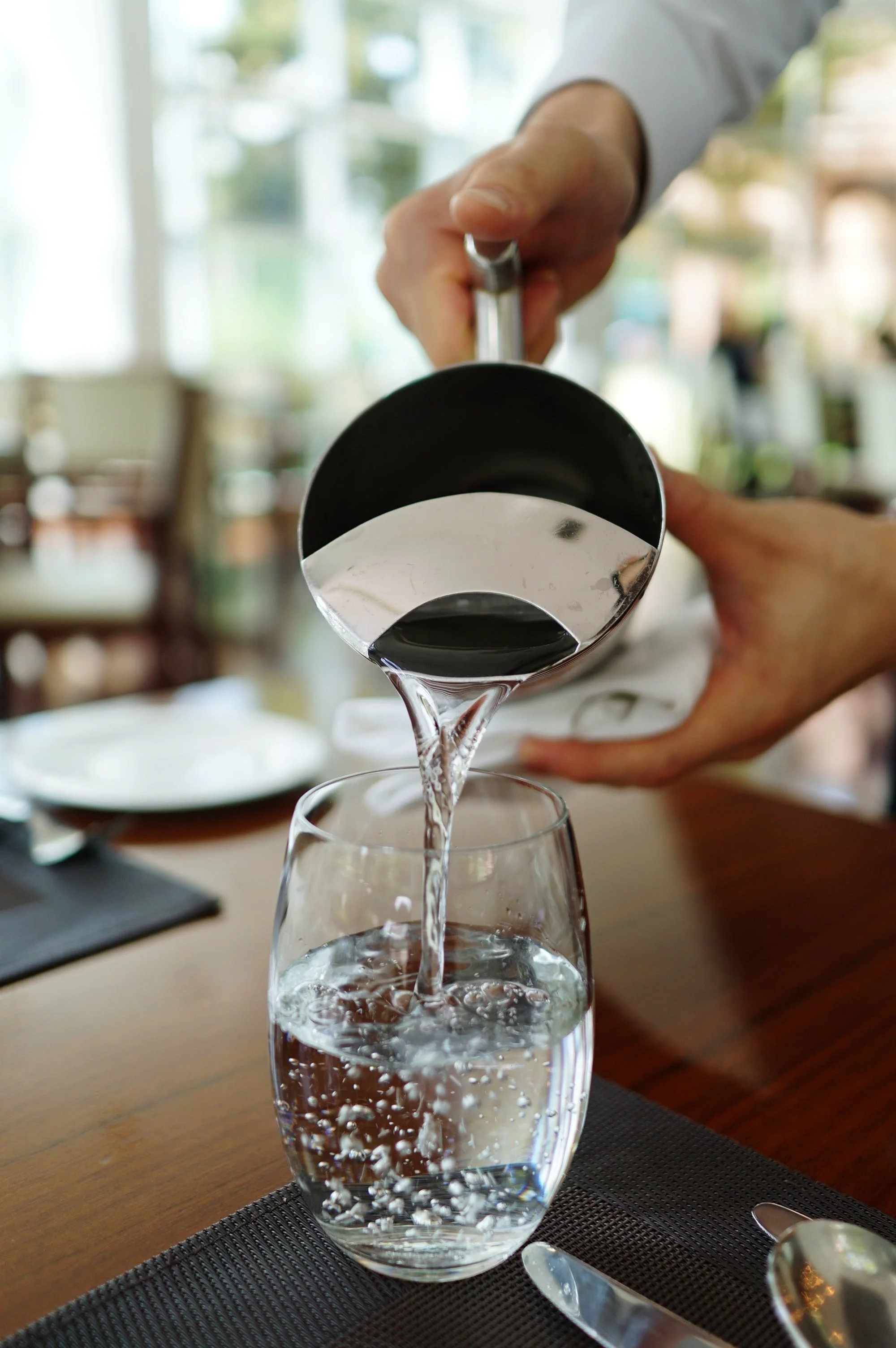 A person pouring water from a metal pitcher into a glass on a wooden dining table.