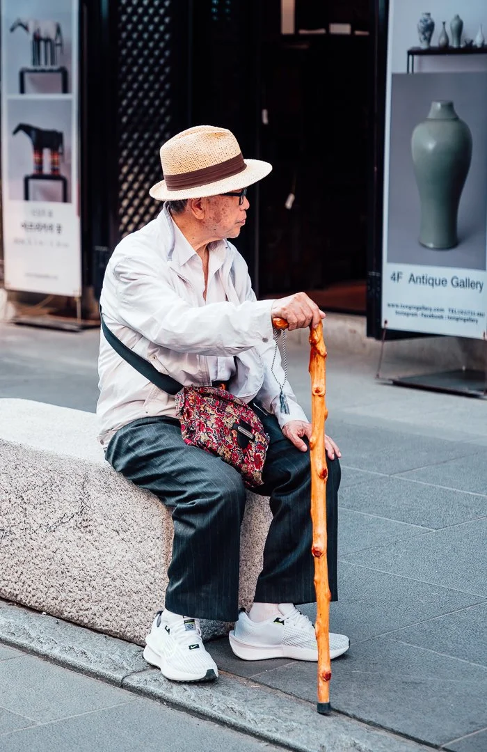 An elderly man wearing a straw hat, glasses, a white long-sleeve shirt, and striped pants sitting on a stone bench on a city sidewalk. He is holding a wooden cane and has a patterned bag slung across his shoulder. Behind him are store signs and displ