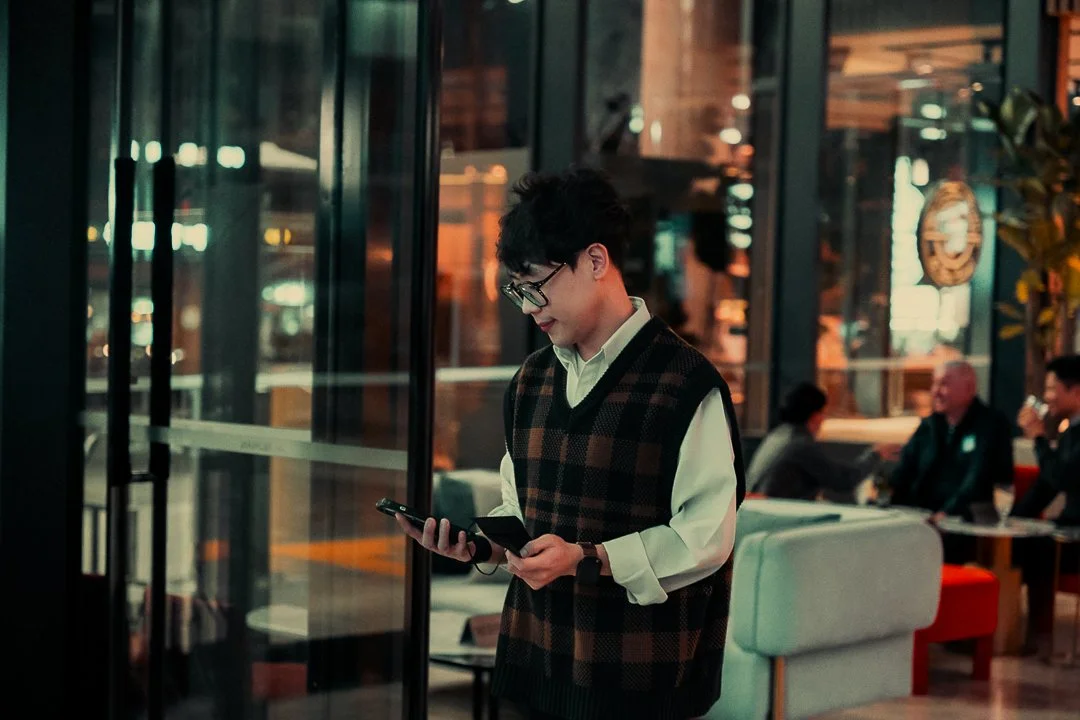 A young man with glasses, dark hair, wearing a checkered vest over a white shirt, standing inside a modern cafe or restaurant, looking at his phone.