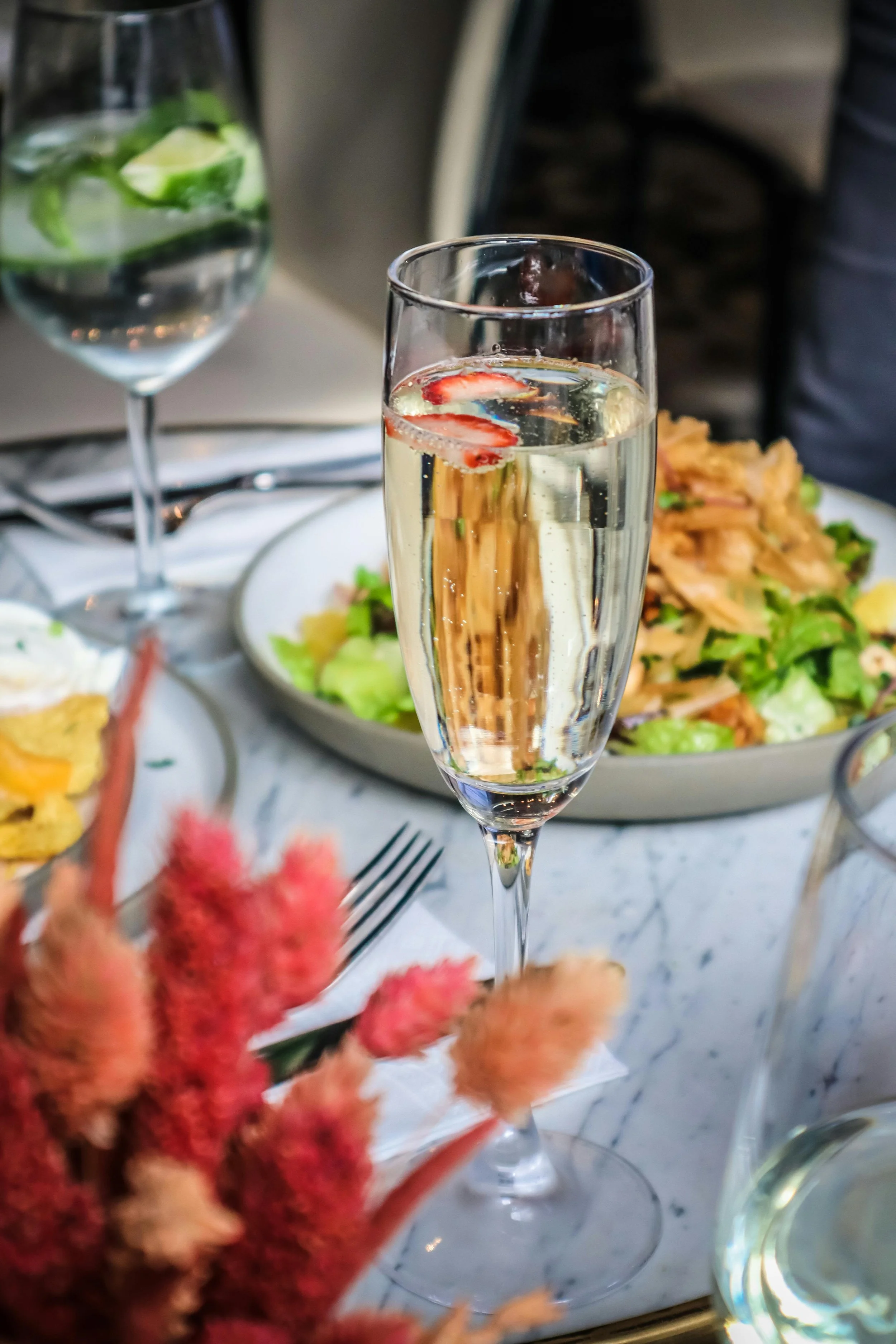 A glass of sparkling wine with strawberries, a glass of water with lime slices, and a bowl of salad on a marble table.
