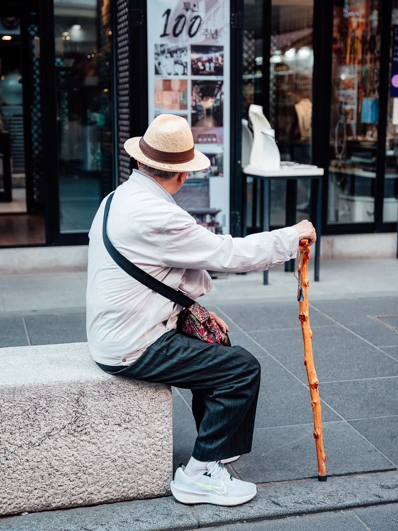 Older man sitting on a stone bench on a city sidewalk, wearing a straw hat, white jacket, black striped pants, and white sneakers, holding a wooden walking stick."