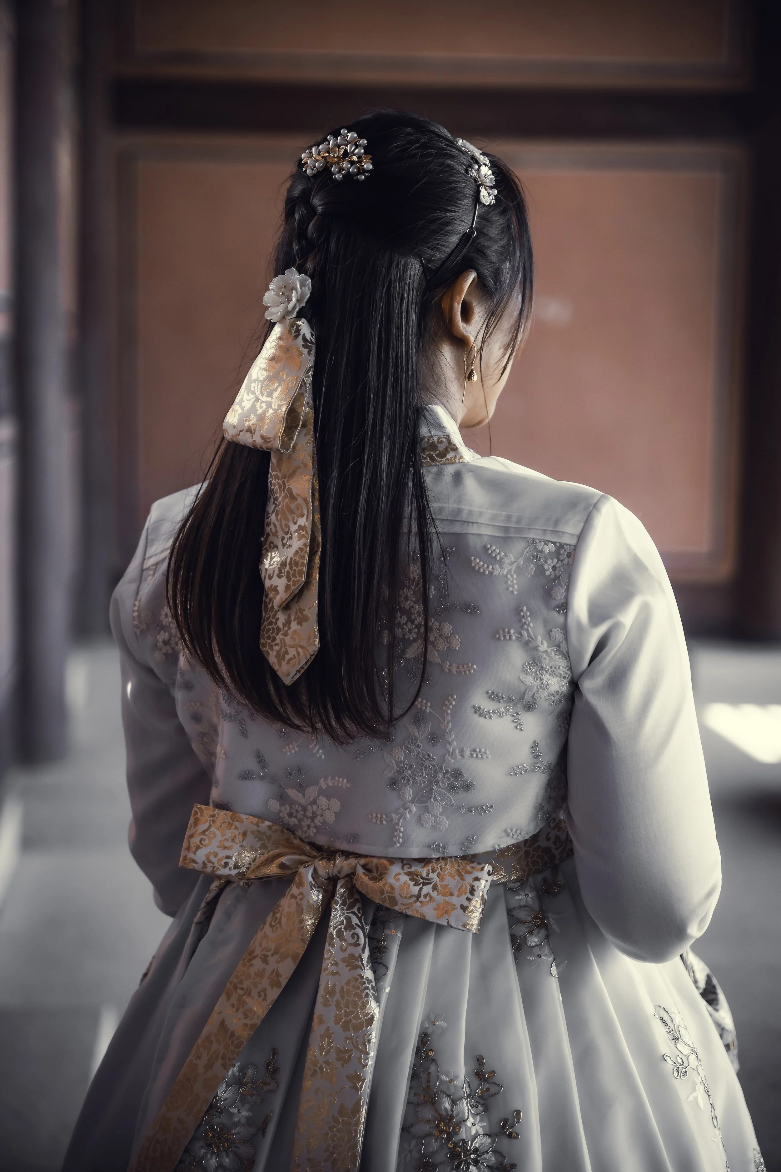 A woman wearing a traditional Korean hanbok with an embroidered jacket and tied bow, adorned with hair accessories including floral pins and a satin ribbon, standing indoors with a red background.