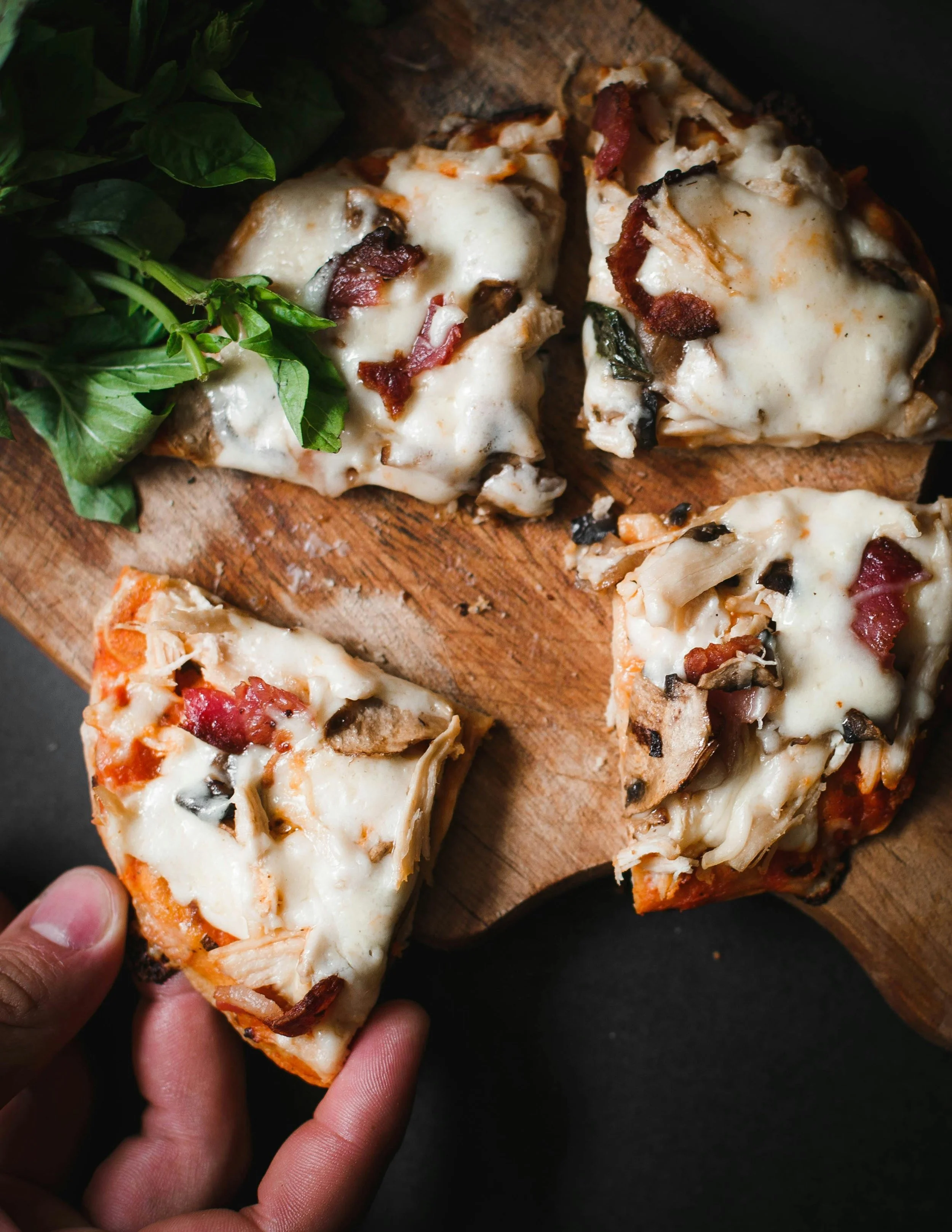 Slices of mushroom, bacon, and cheese pizza on a wooden cutting board, with fresh basil on the side.