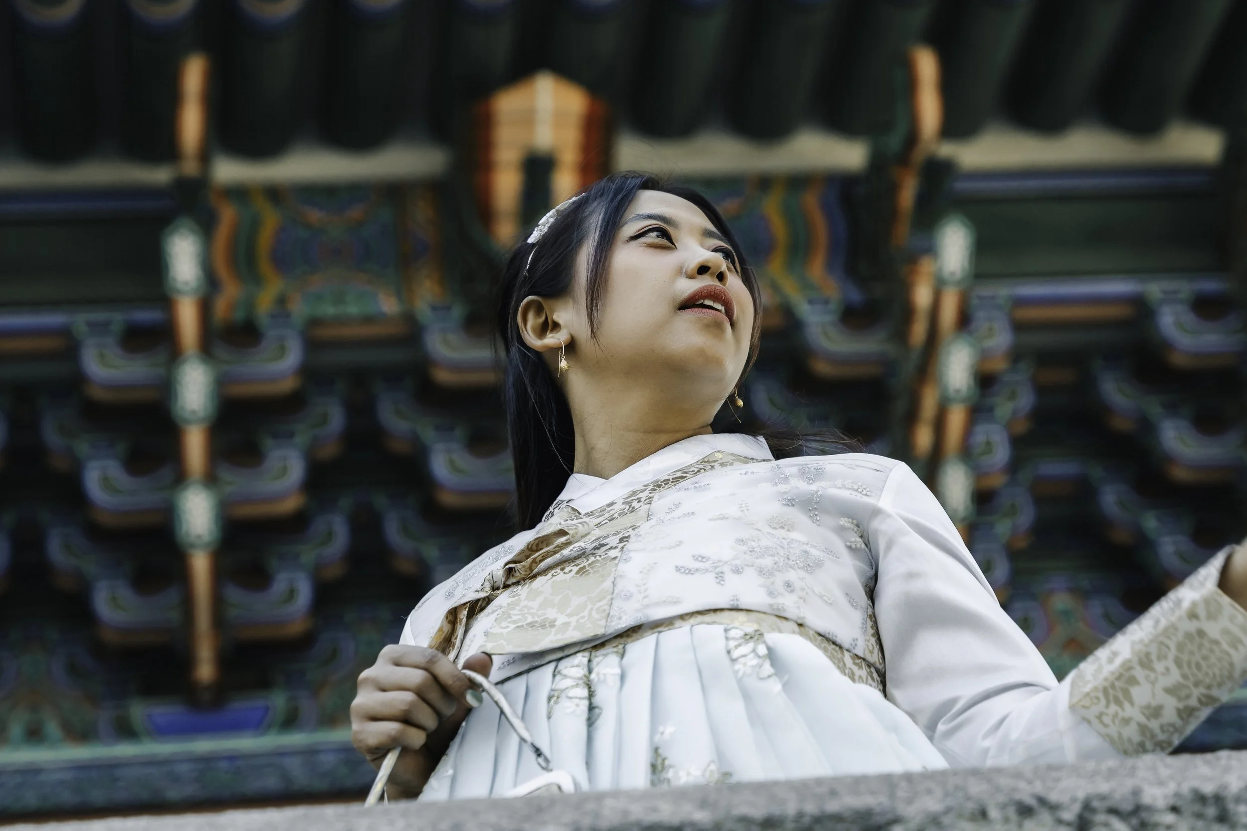Young woman in traditional Korean hanbok dress looking upwards, standing in front of a colorful, ornate building with intricate patterns.