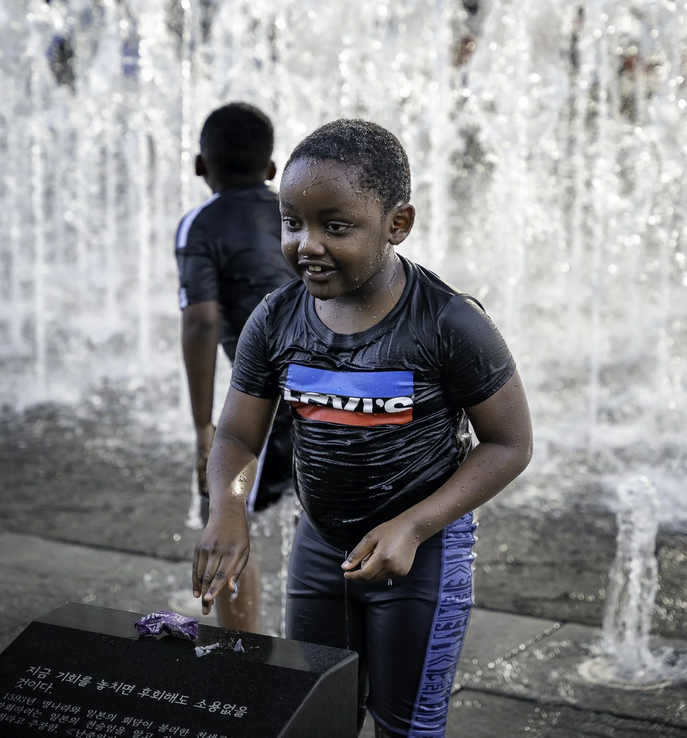A young boy in a black Levi's shirt plays in a fountain, getting soaked with water falling around him, with another child in the background.