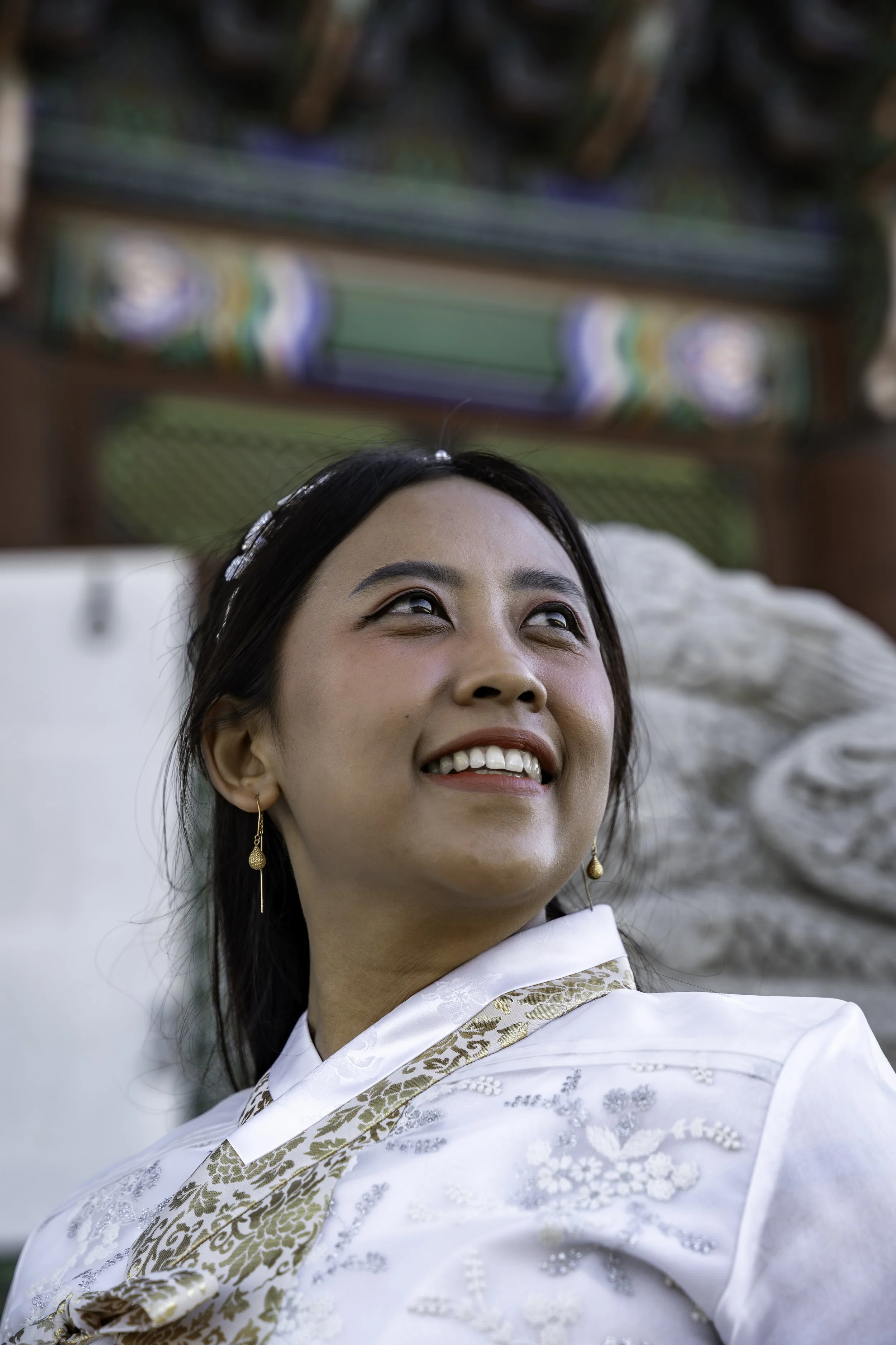 A smiling woman wearing traditional Asian attire, standing outdoors near a building with colorful decorative elements and a sculpted stone lion.