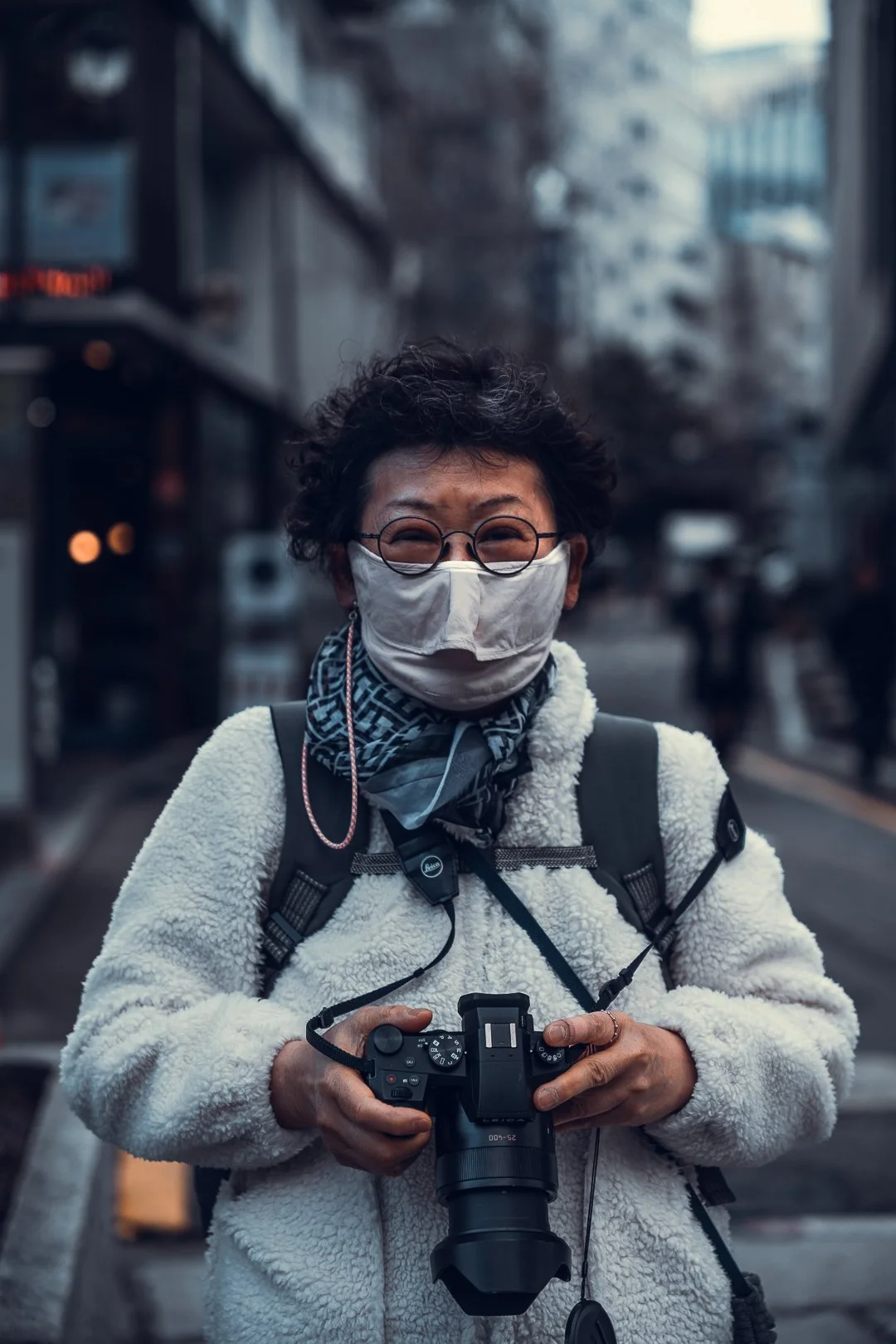 Woman wearing glasses, a face mask, and a white fleece jacket, holding a camera on a city street.