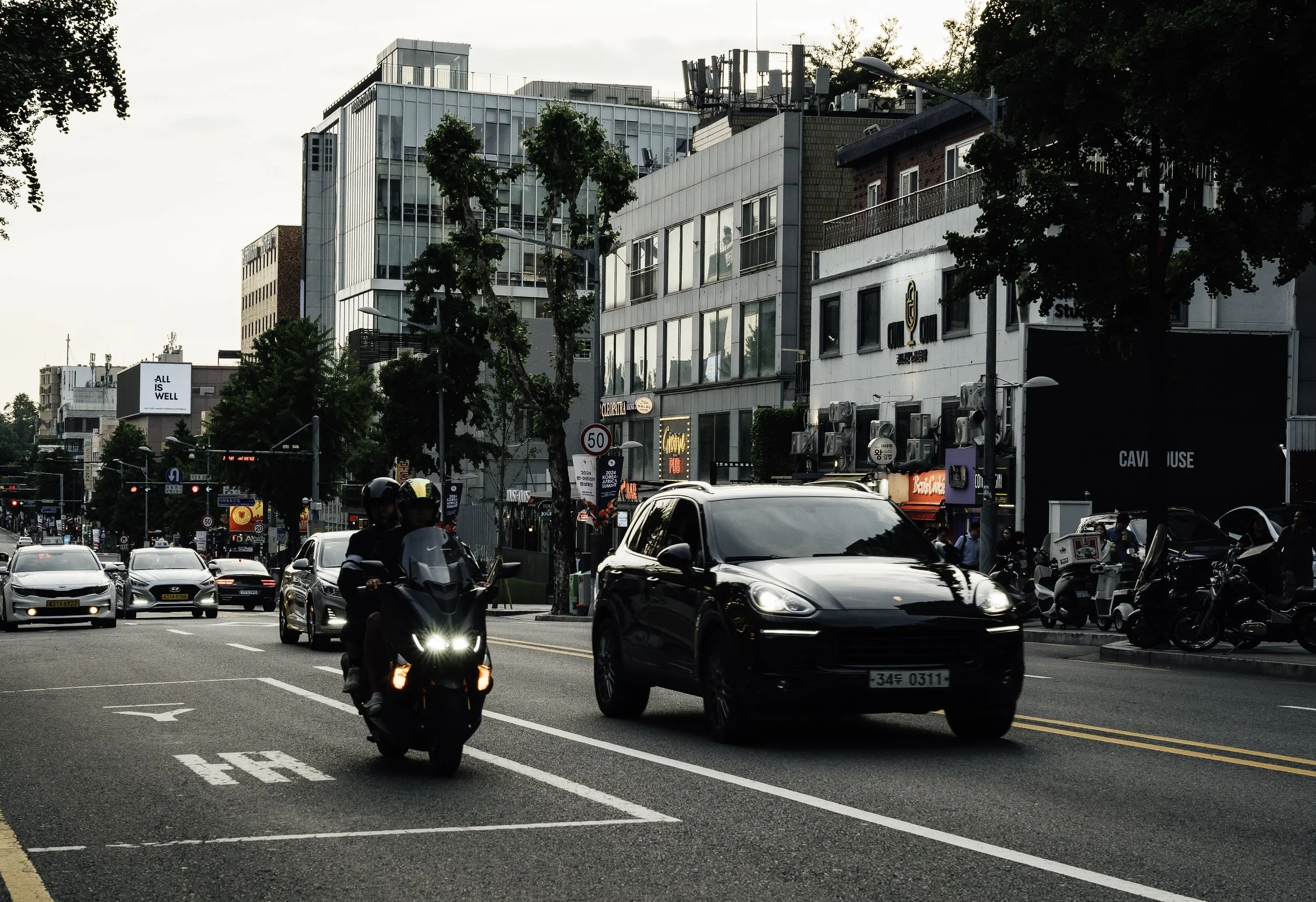 Urban street scene with cars and motorcycles, modern buildings, trees, and city signs in the evening