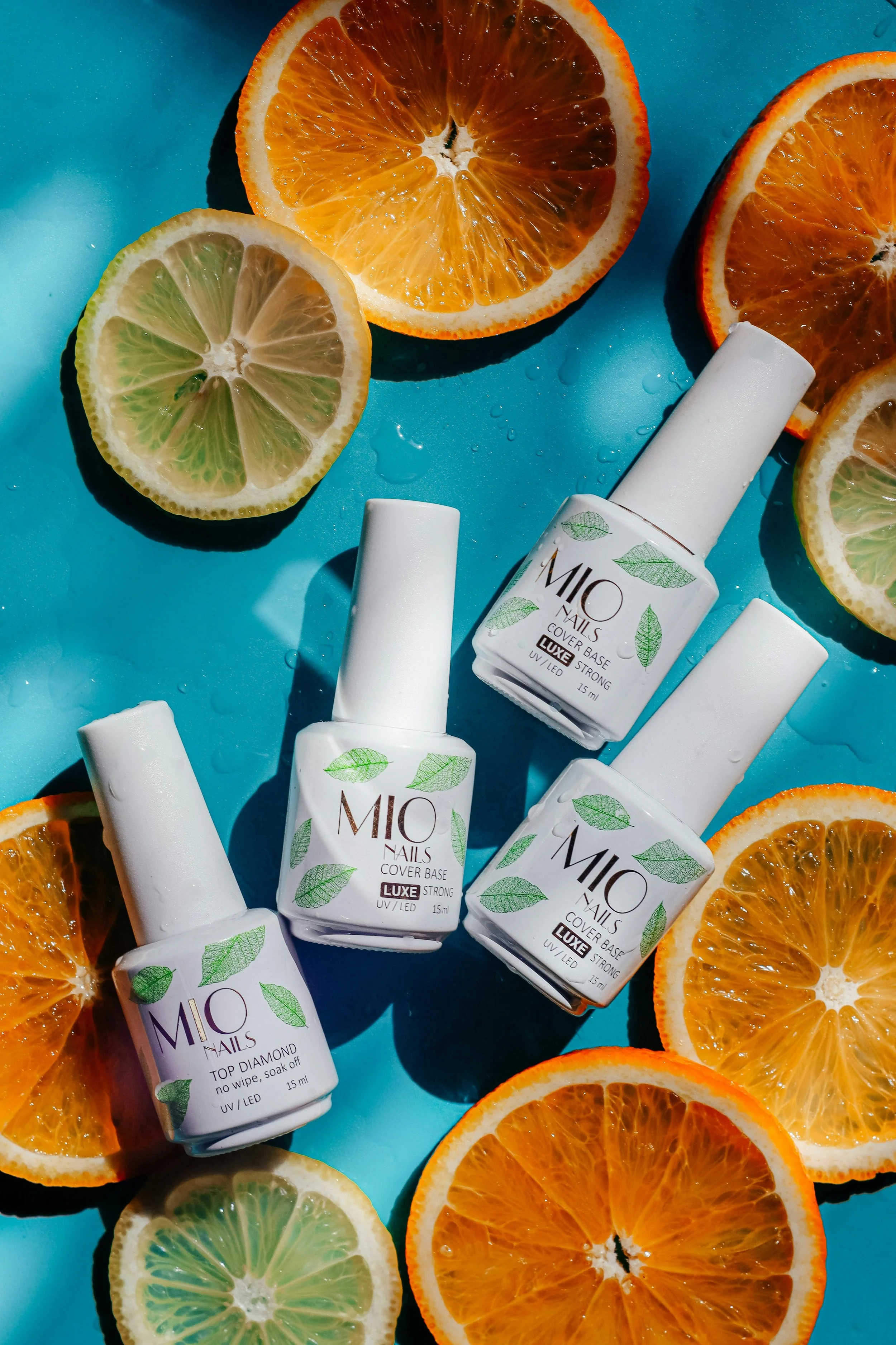 Several bottles of MIO nail cover base and top diamond, surrounded by slices of orange and lemon, on a blue background with water droplets.