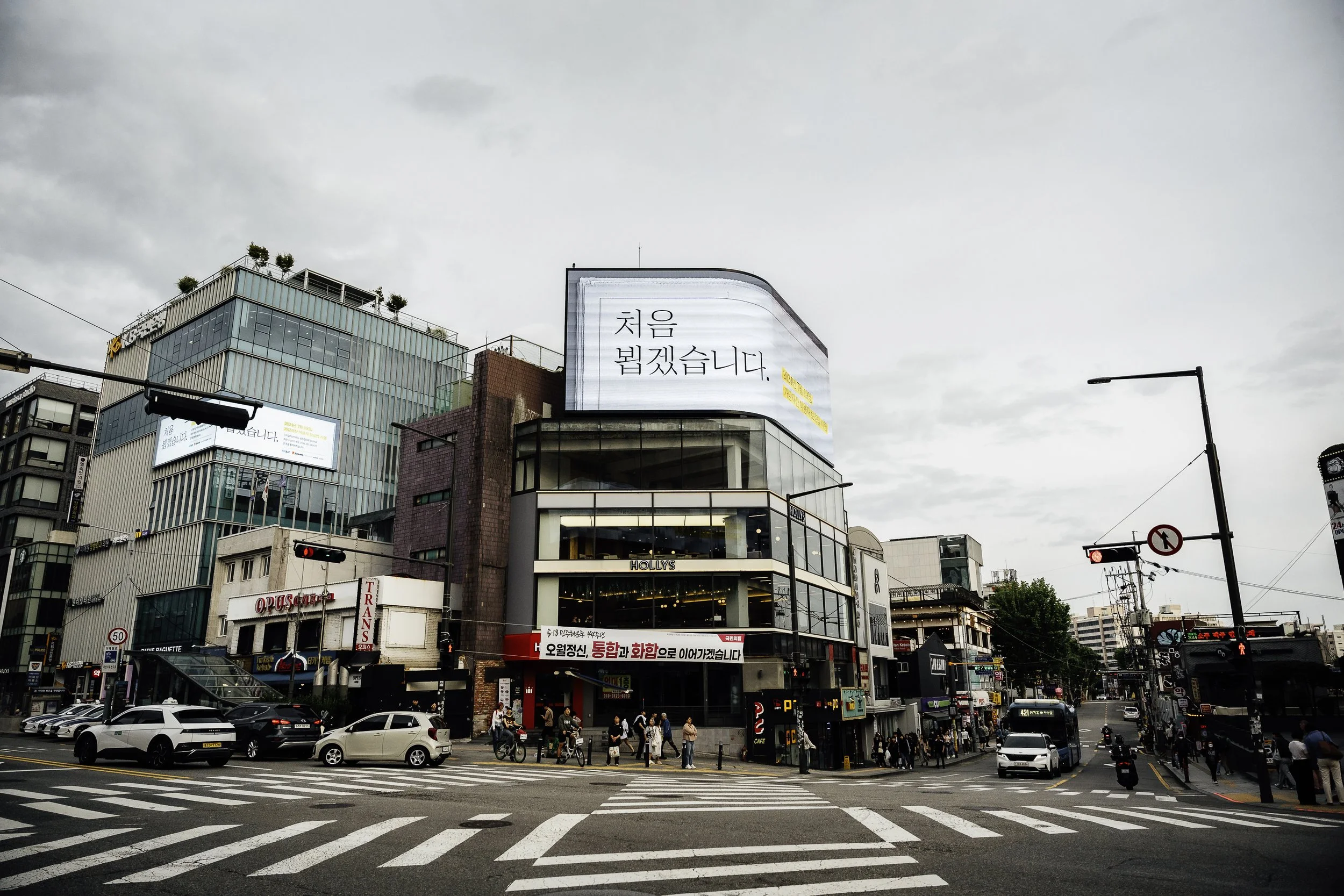 Urban street scene with cars, pedestrians, and buildings, including a modern corner building with a curved glass facade and electronic billboards, and traffic lights showing red in a city.