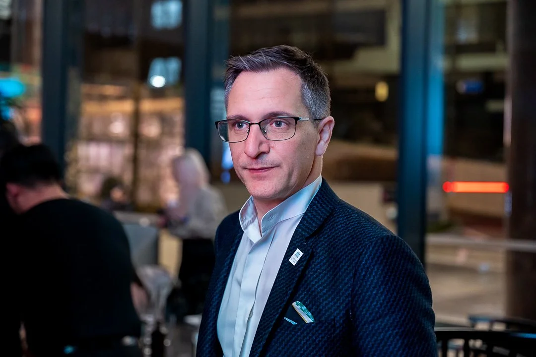 A man with glasses in a dark blazer and light-colored shirt standing indoors at a social gathering.