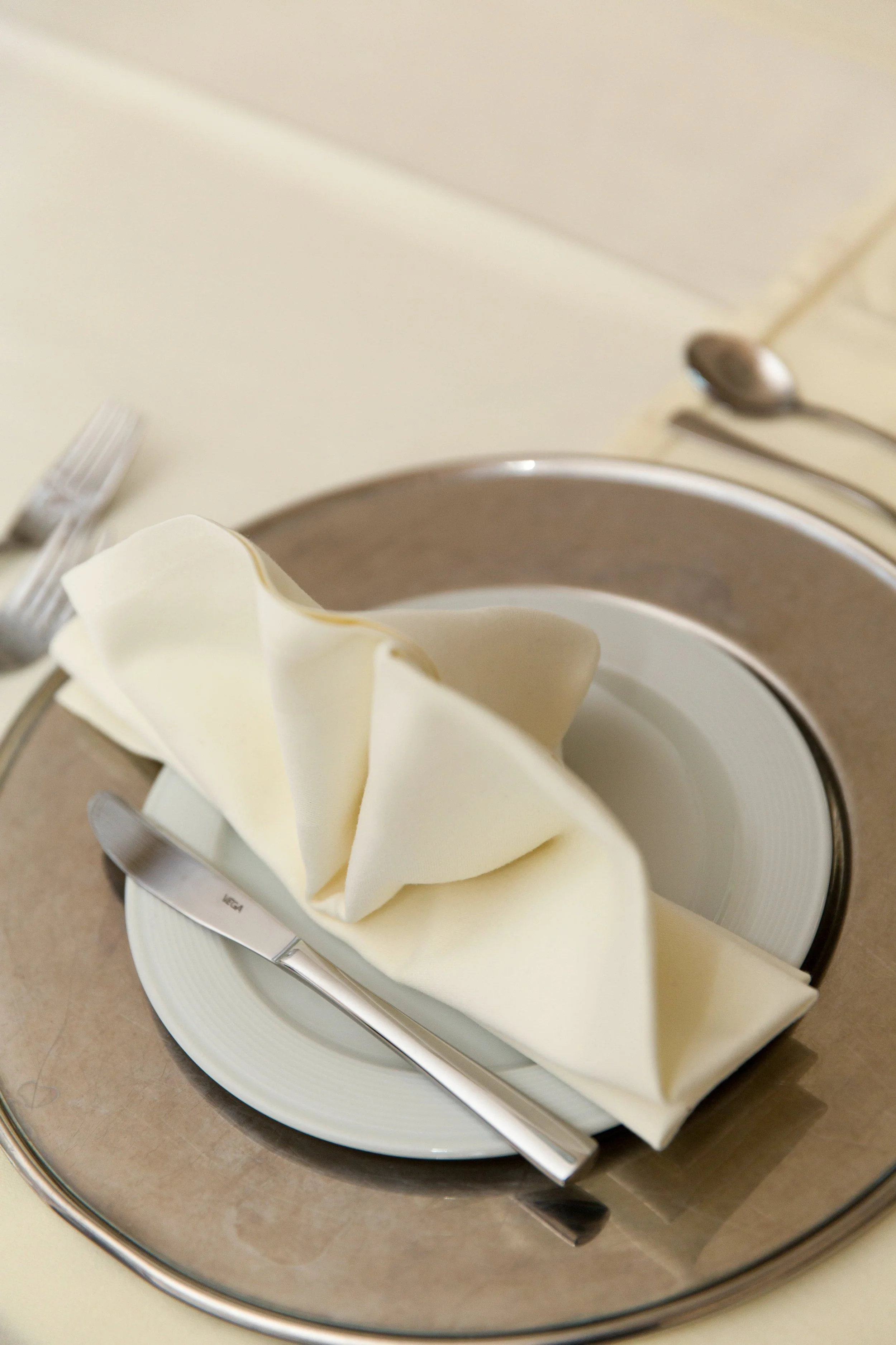 A place setting with a white plate, a folded cloth napkin, a butter knife, and a fork, on a beige tablecloth. A silver serving tray surrounds the plate.