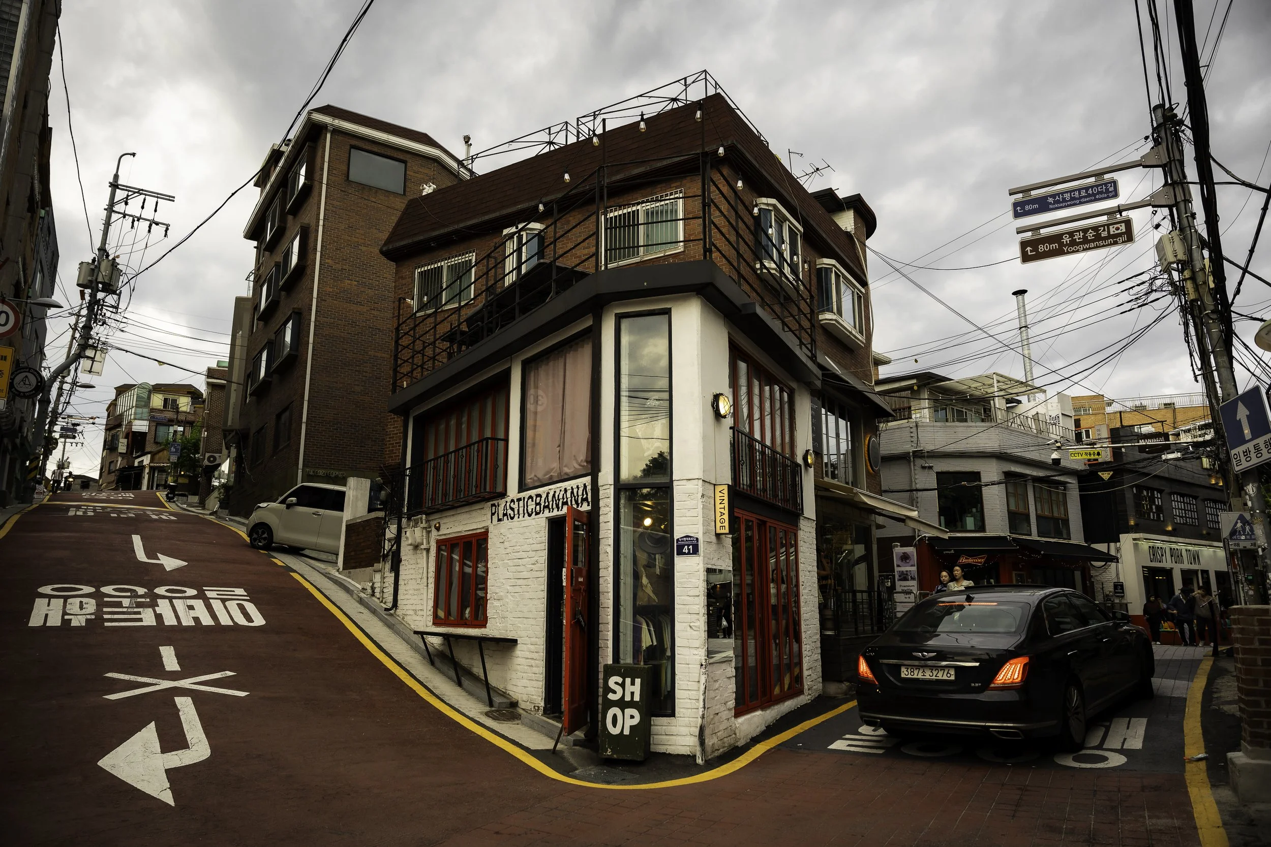 A corner building with red framed windows and white brick facade, hosting a shop called 'Plastic Banana,' on a narrow street with parked cars, overhead power lines, and surrounding buildings, under a cloudy sky.