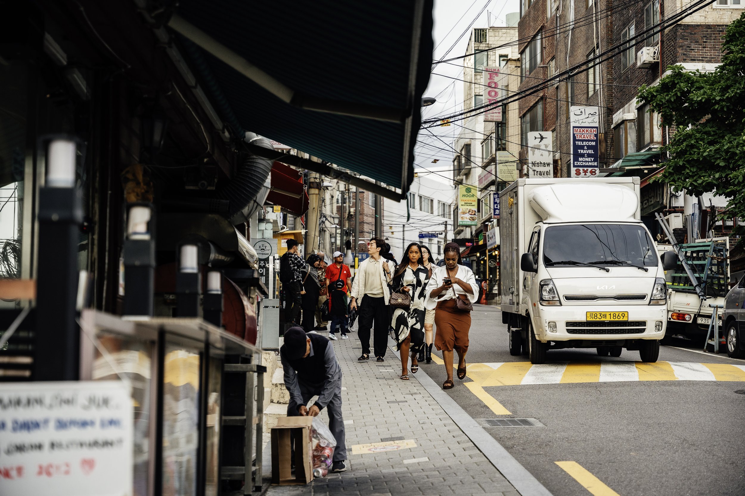 Busy city street with pedestrians, parked cars, a white delivery truck, and various storefronts with signs in multiple languages.