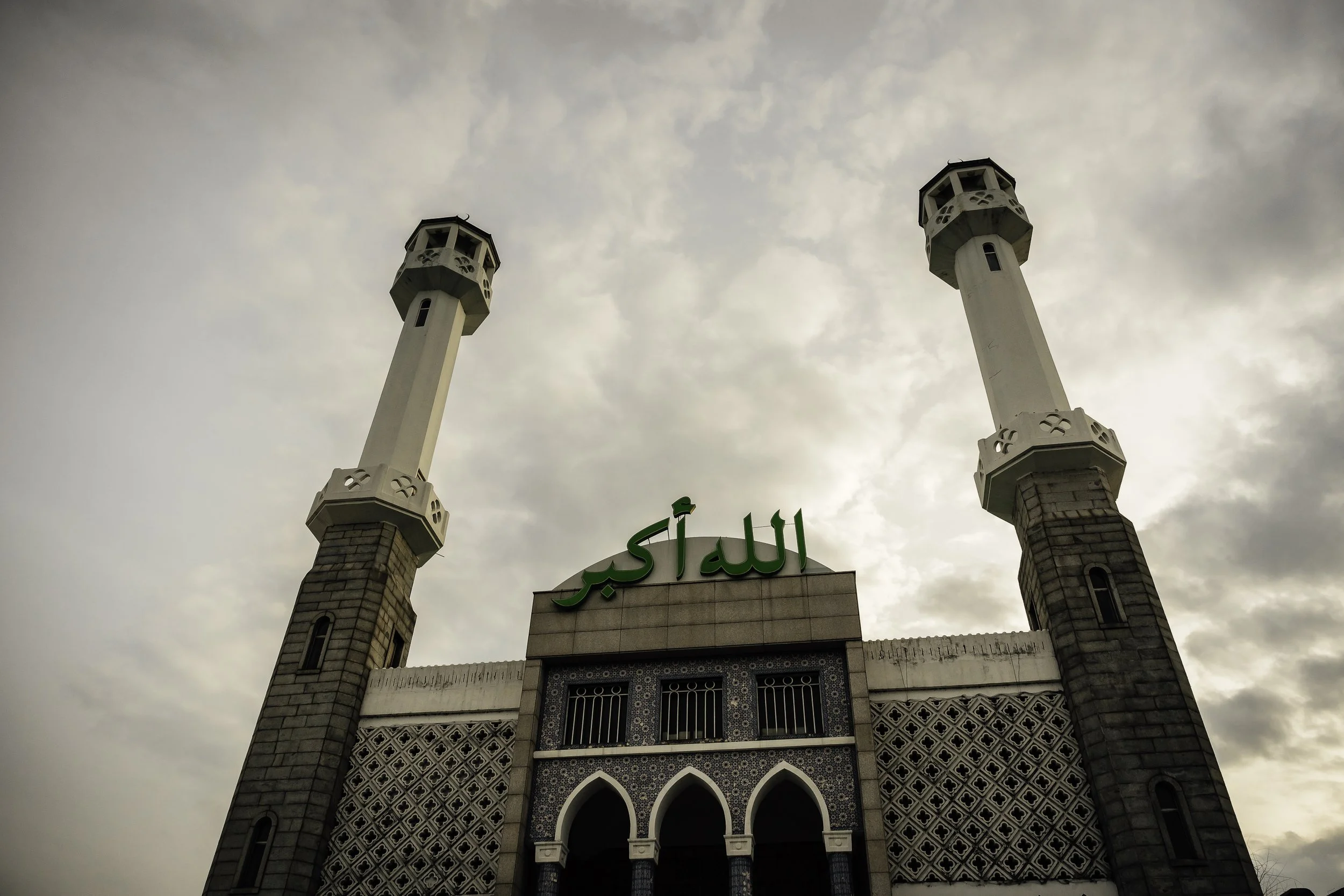 Front view of a mosque with two minarets and green Arabic script on the facade, under cloudy sky.