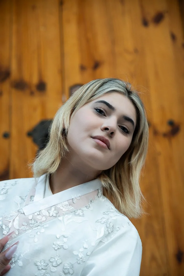 Young woman with blonde hair in a white embroidered top standing in front of a wooden wall.