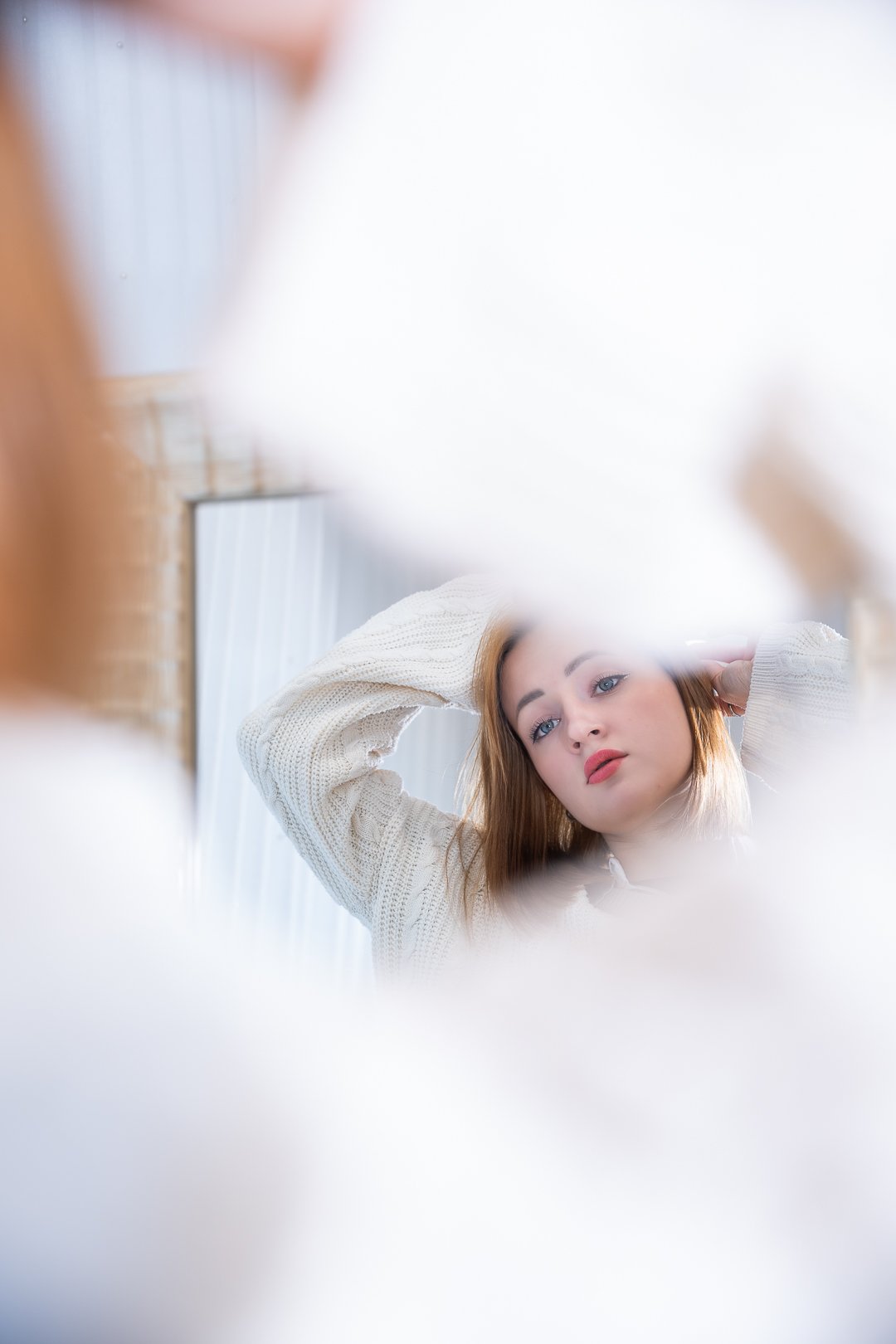 A young woman with red hair looking into a mirror, adjusting her hair, with her face reflected in the mirror.