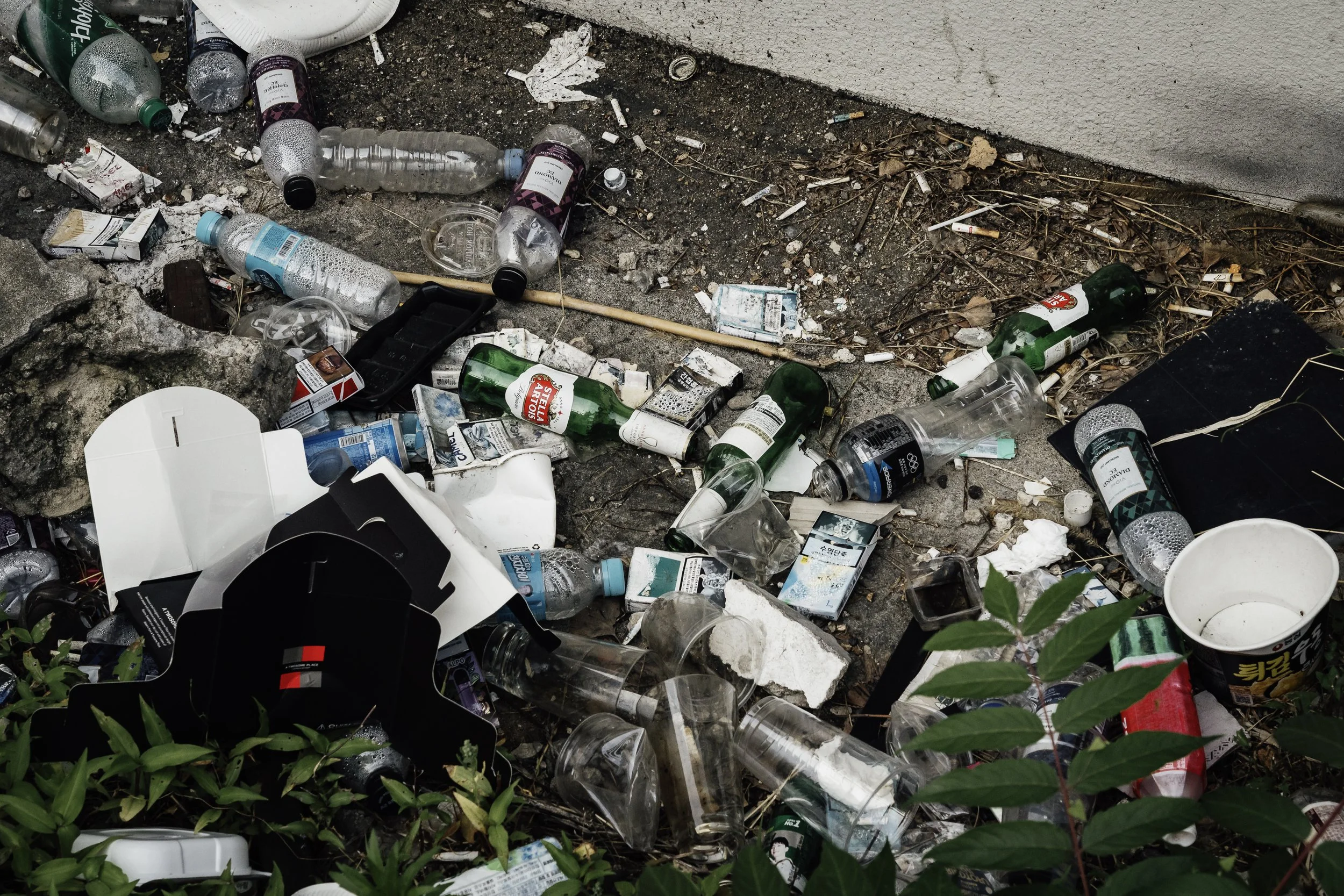 Littered trash on the ground including plastic bottles, cups, cigarette packs, and paper debris near a wall and some green plants.