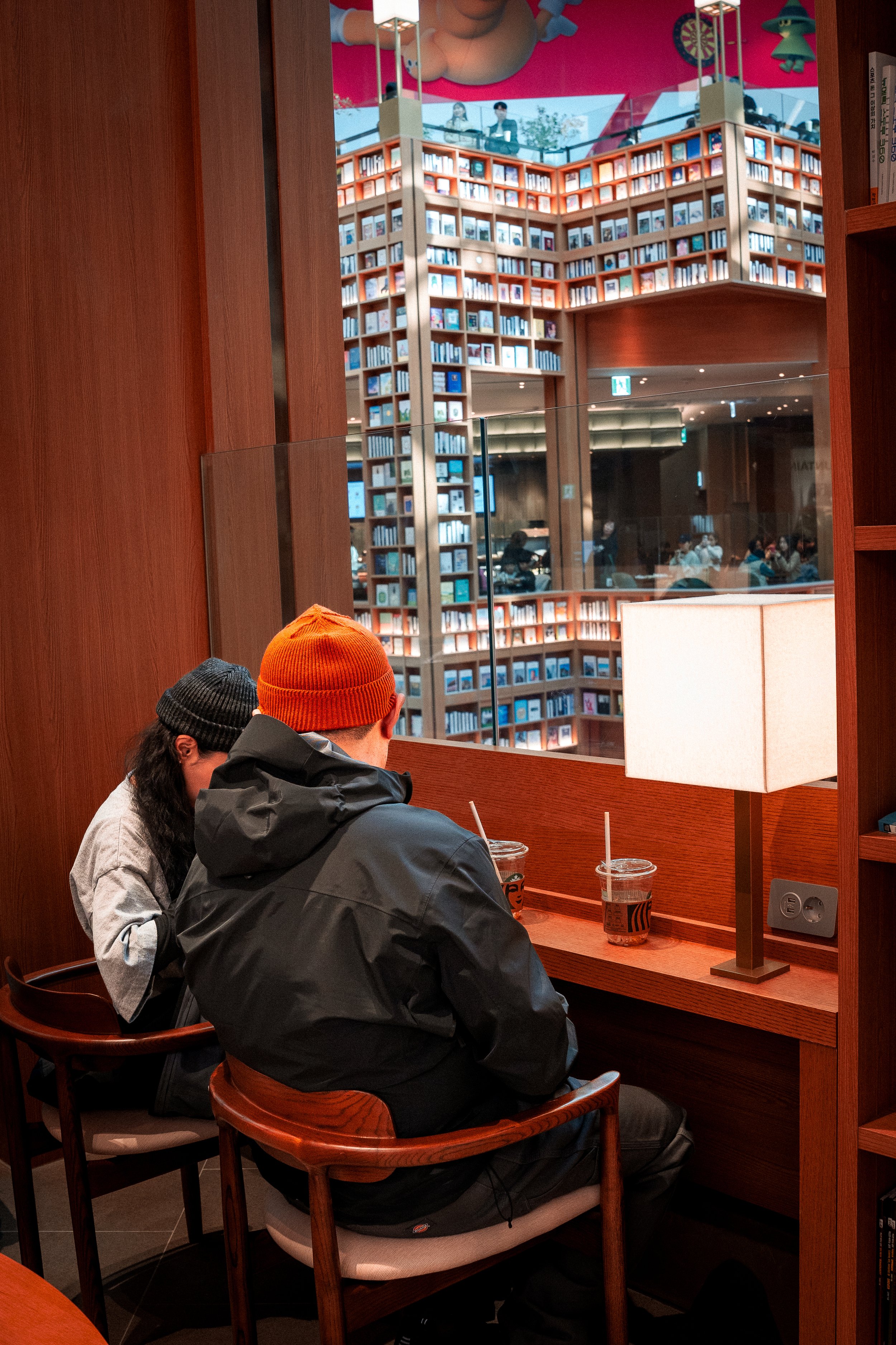 Two people sitting in a cozy coffee shop, viewed through a large wooden framed window, with a large bookshelf in the background.
