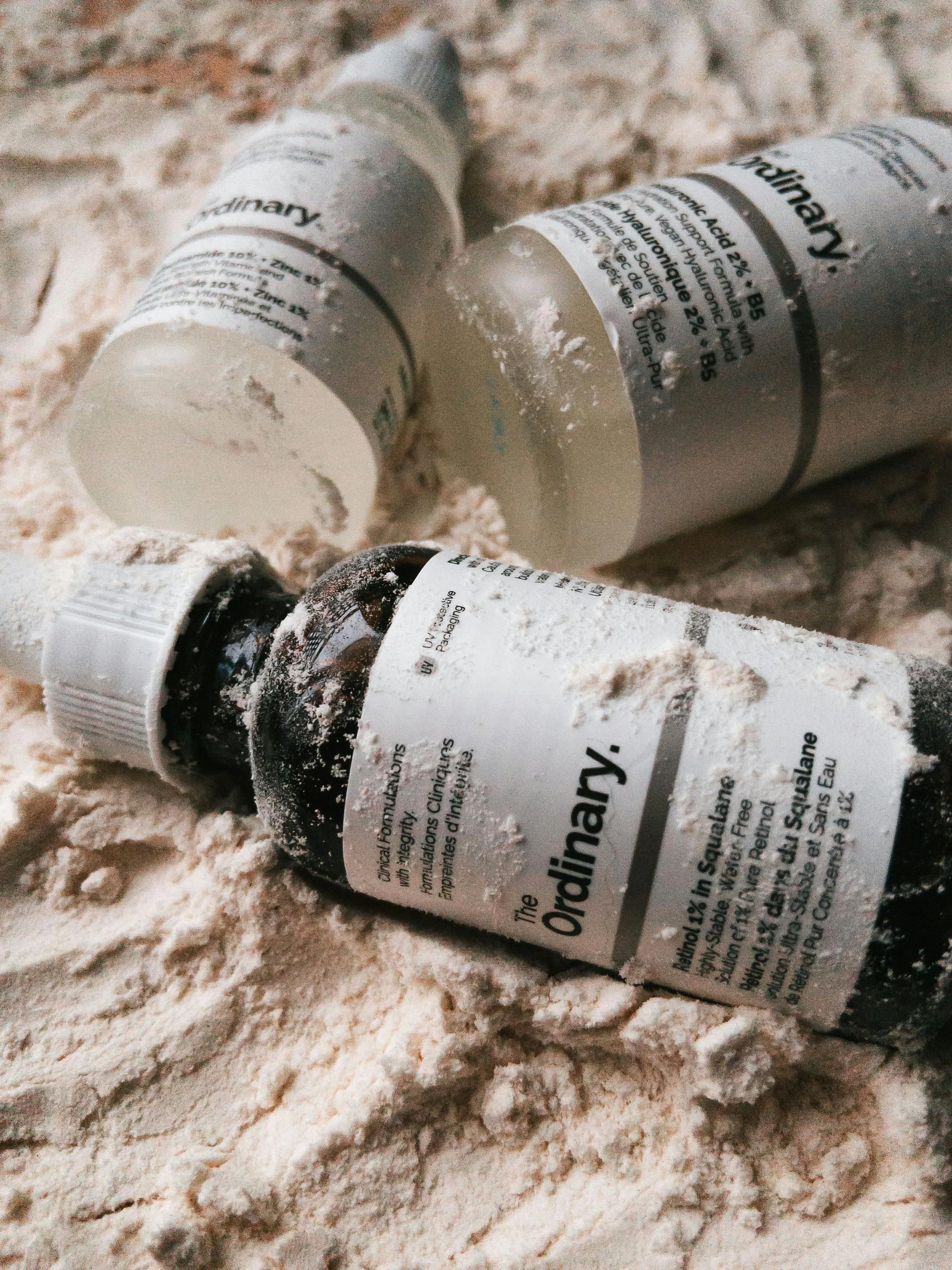 Three pill bottles labeled 'The Ordinary' lying on a sandy surface, with the bottles partially covered in sand and dust.