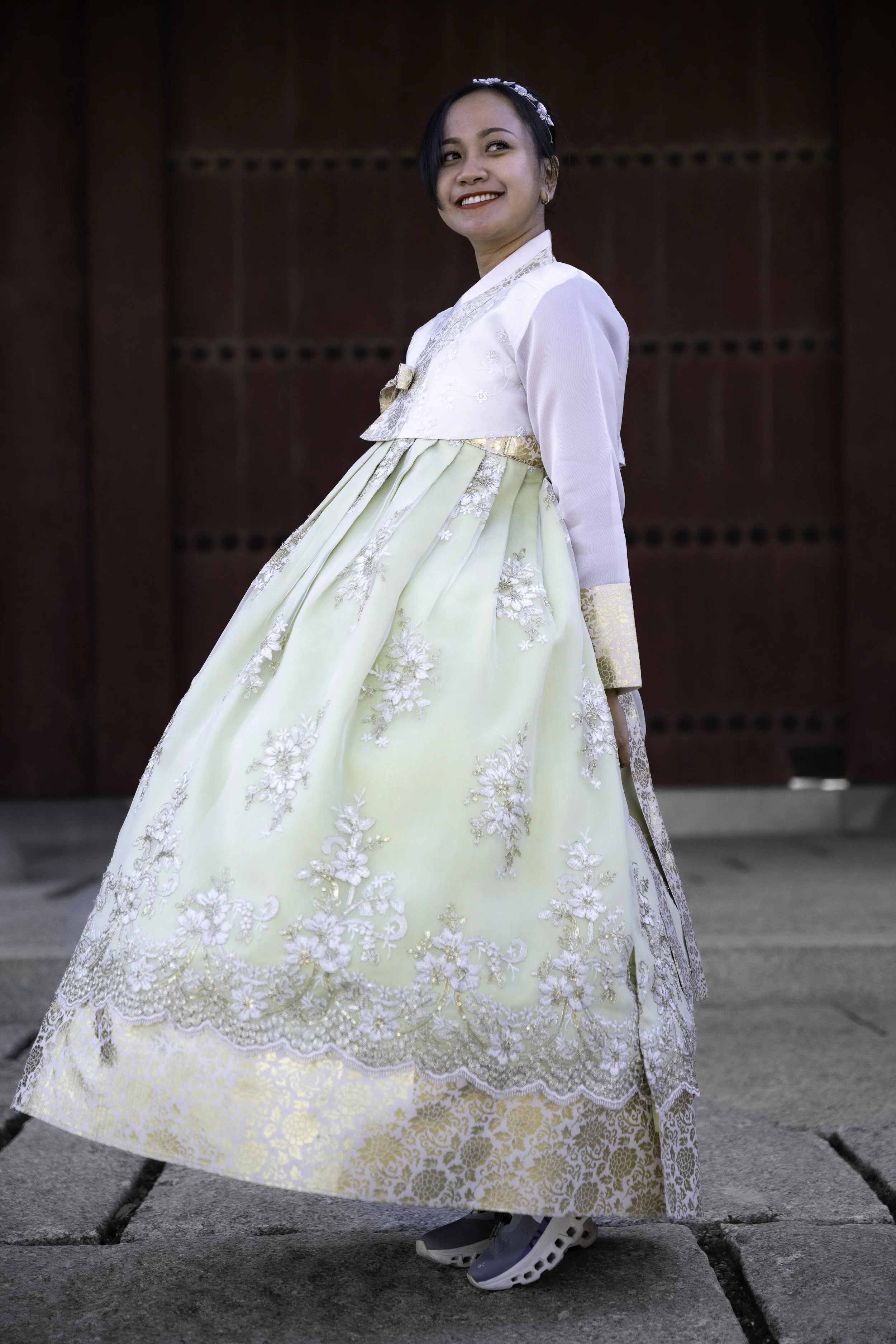 Young woman smiling, wearing a traditional Korean hanbok dress with a pale yellow skirt and white top, standing outdoors on stone pavement.
