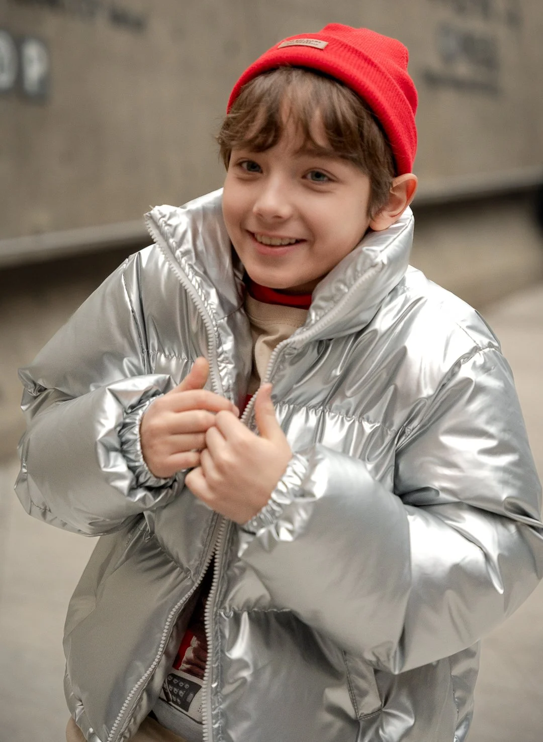 A young boy smiling, wearing a red beanie, a shiny silver puffer jacket, and casual clothes, in an indoor setting.