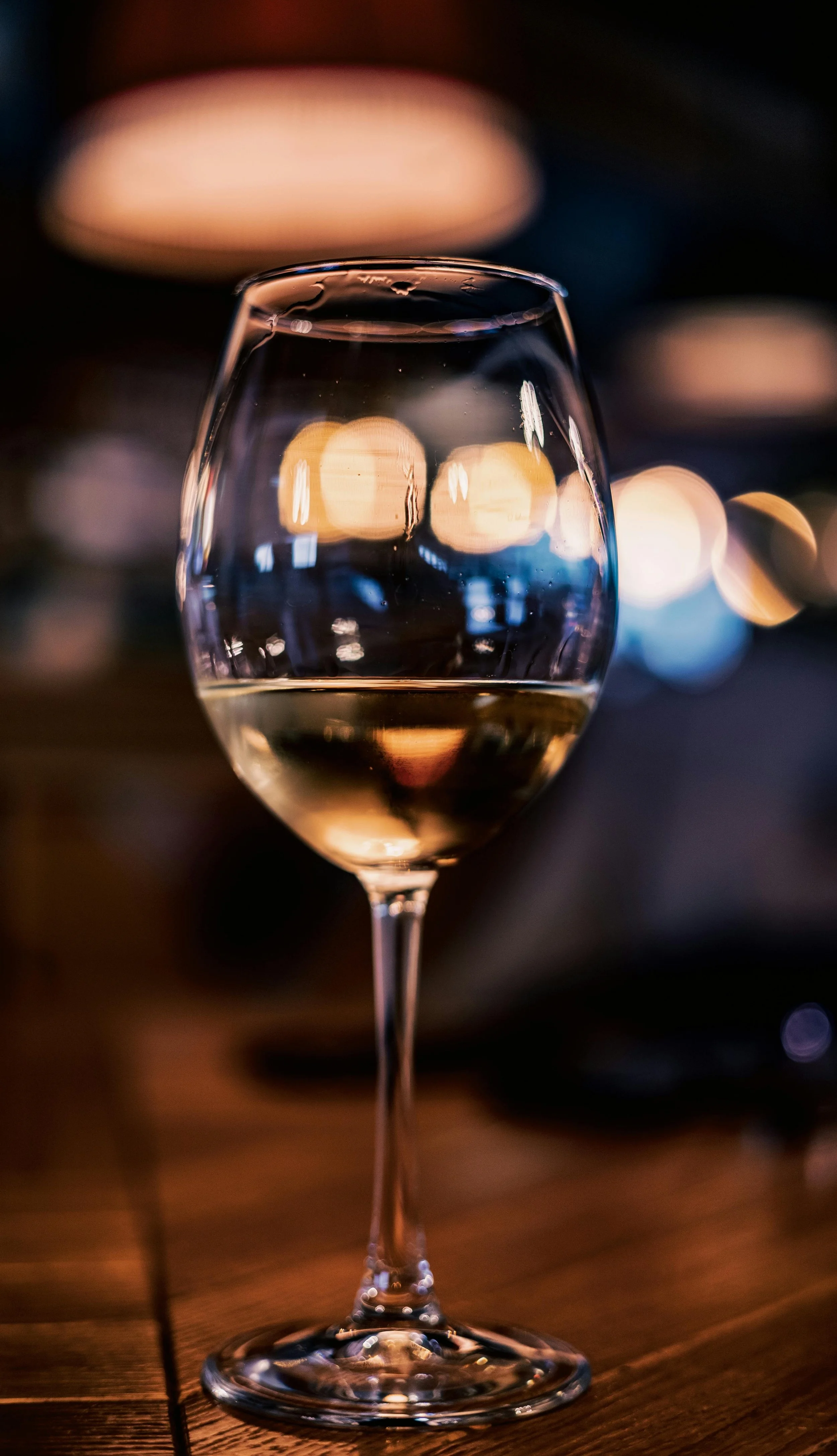 A glass of white wine on a wooden table with blurred bokeh lights in the background.