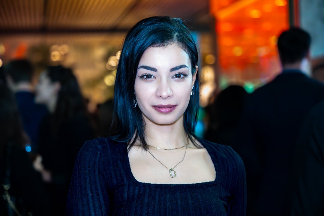 A young woman with dark hair and makeup at a social event, wearing a black top, gold necklace, and earrings, with blurred people in the background.