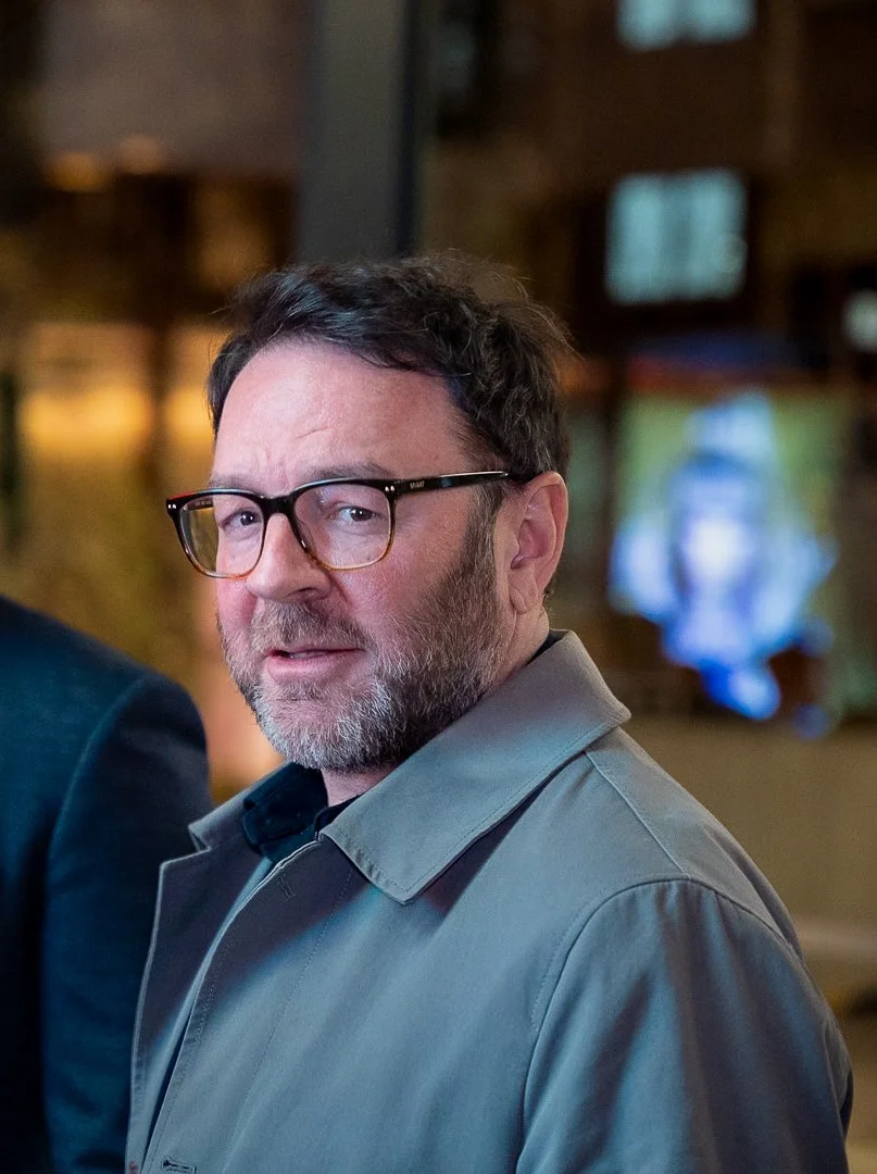 A middle-aged man with dark hair and glasses, wearing a gray jacket, looking at the camera in an indoor setting.
