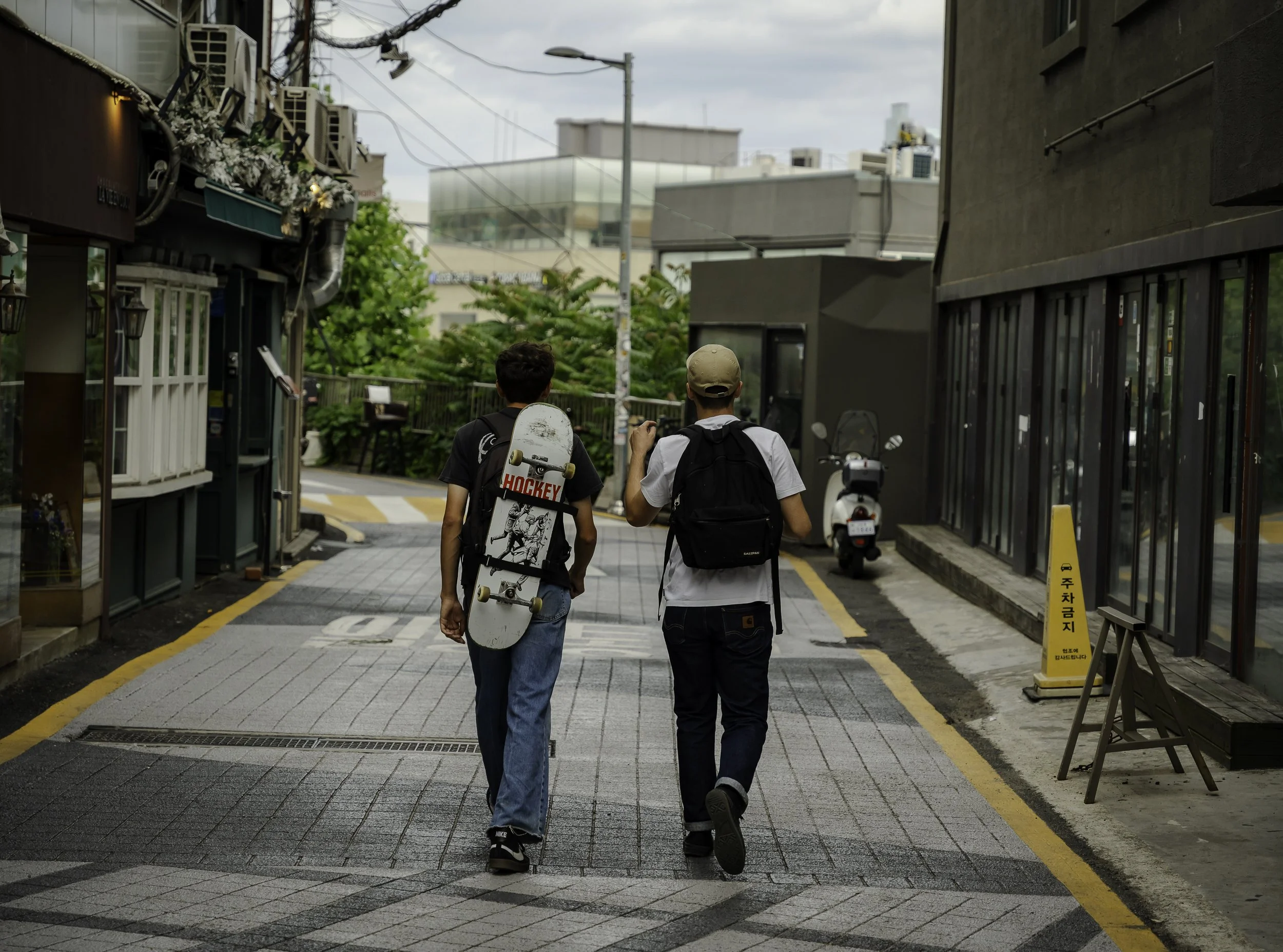 Two young men walking down a city street with one carrying a skateboard on his shoulder. The street has modern buildings and is lined with trees and parked scooter.