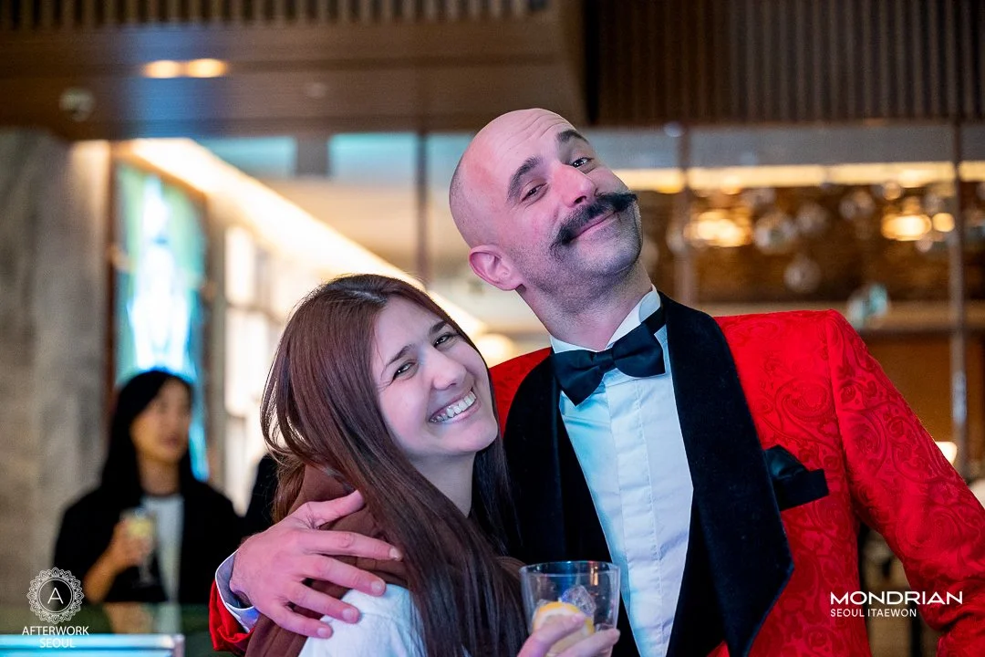 A man with a mustache wearing a tuxedo with a red patterned jacket and black bow tie hugs a woman with long brown hair, smiling. The woman holds a glass. In the background, a woman in black is holding a drink.
