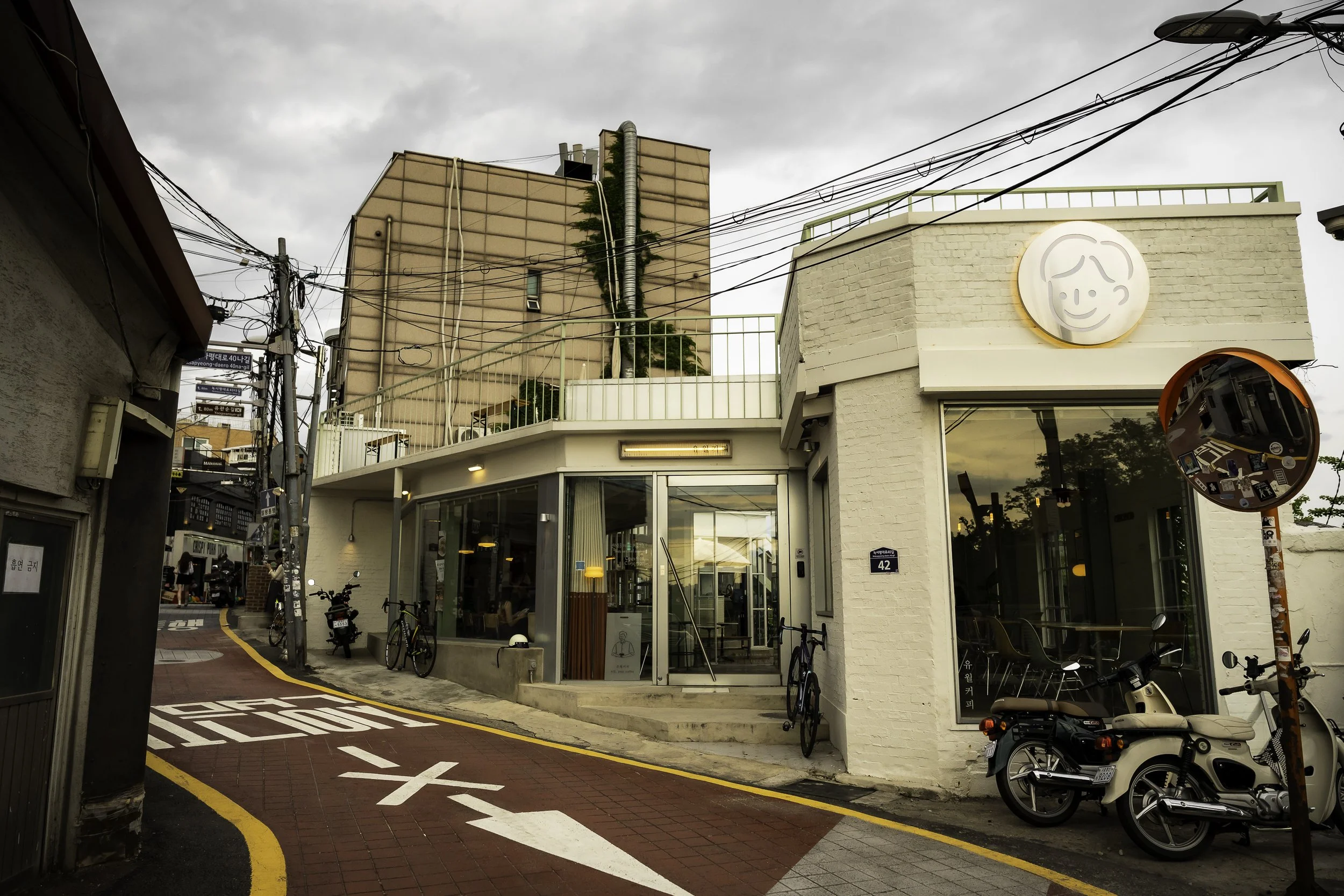 A corner cafe with large glass windows, a white brick exterior, and an illuminated sign of a smiling face logo. There are bikes and scooters parked outside, a traffic mirror on a pole, and a narrow street with markings and a curve.