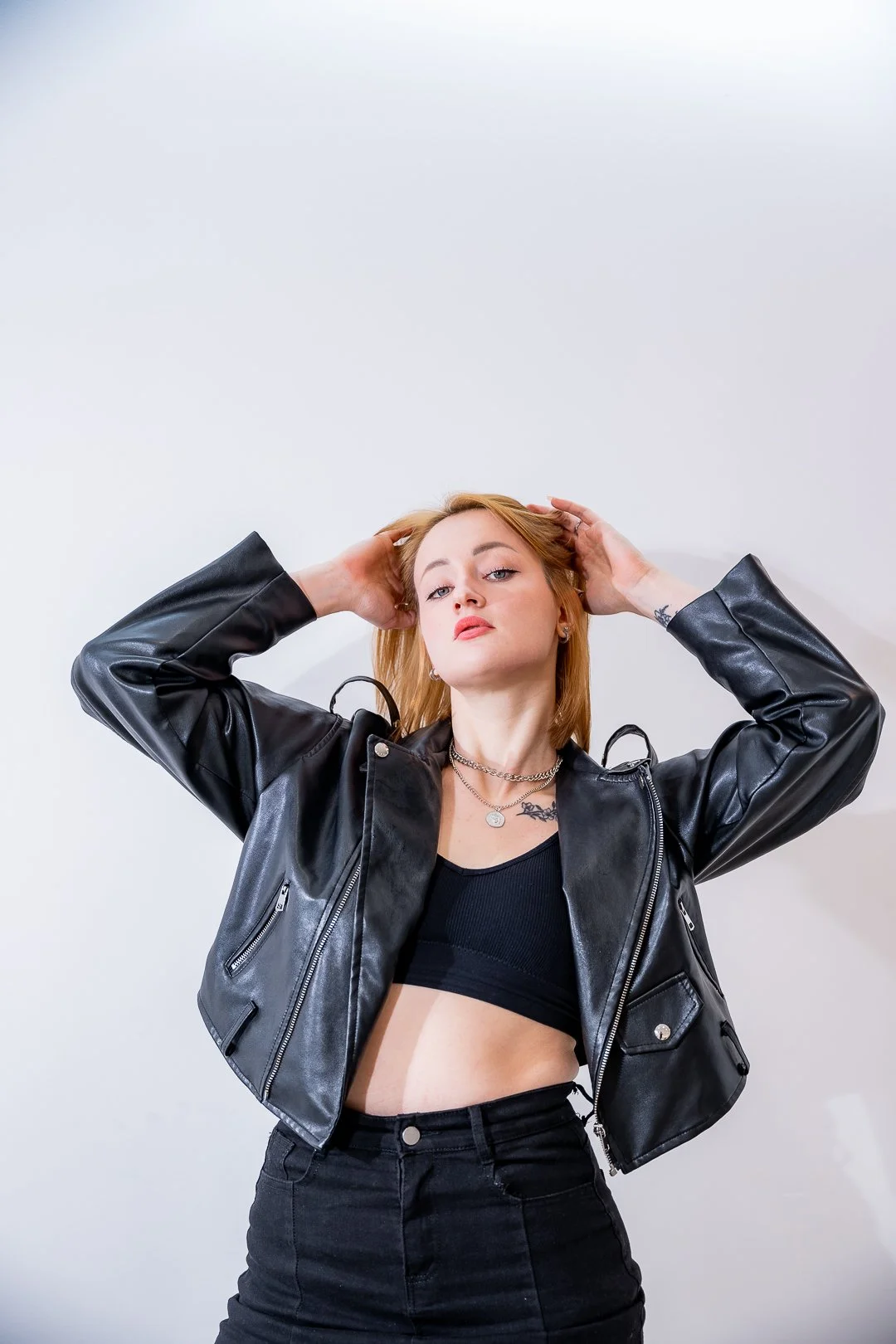 A young woman with blonde hair wearing a black leather jacket, black crop top, and black high-waisted pants; posing with her hands behind her head against a plain white background.