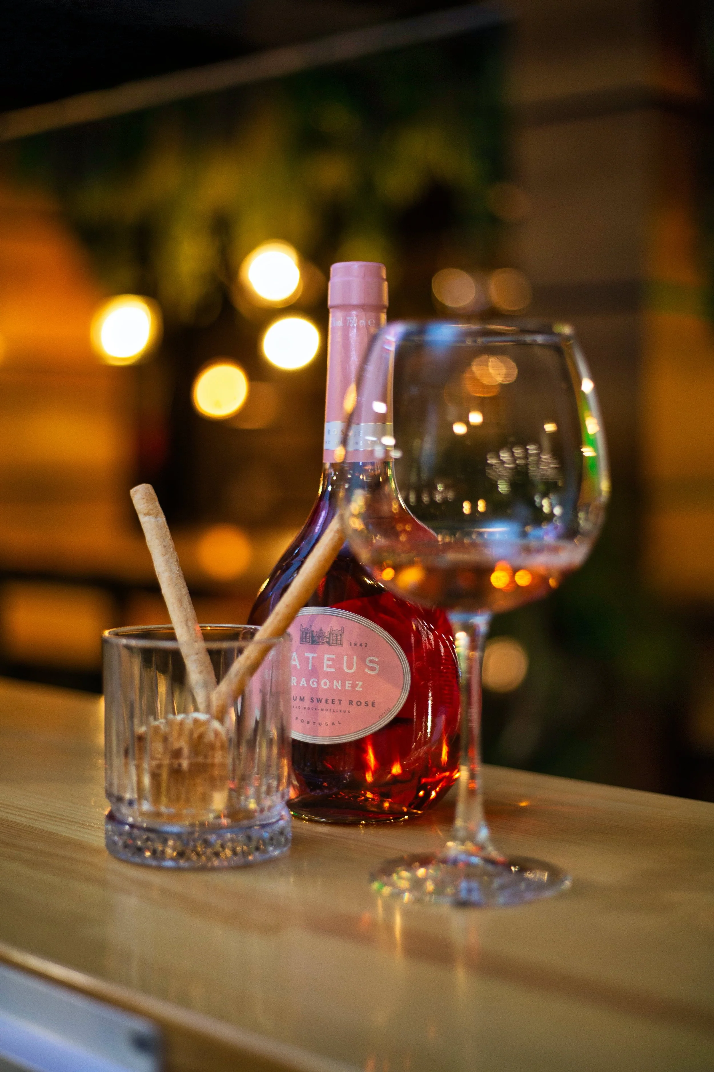 A bottle of rosé wine, a glass of rosé wine, and a small glass with breadsticks on a wooden table, with blurred warm lights in the background.
