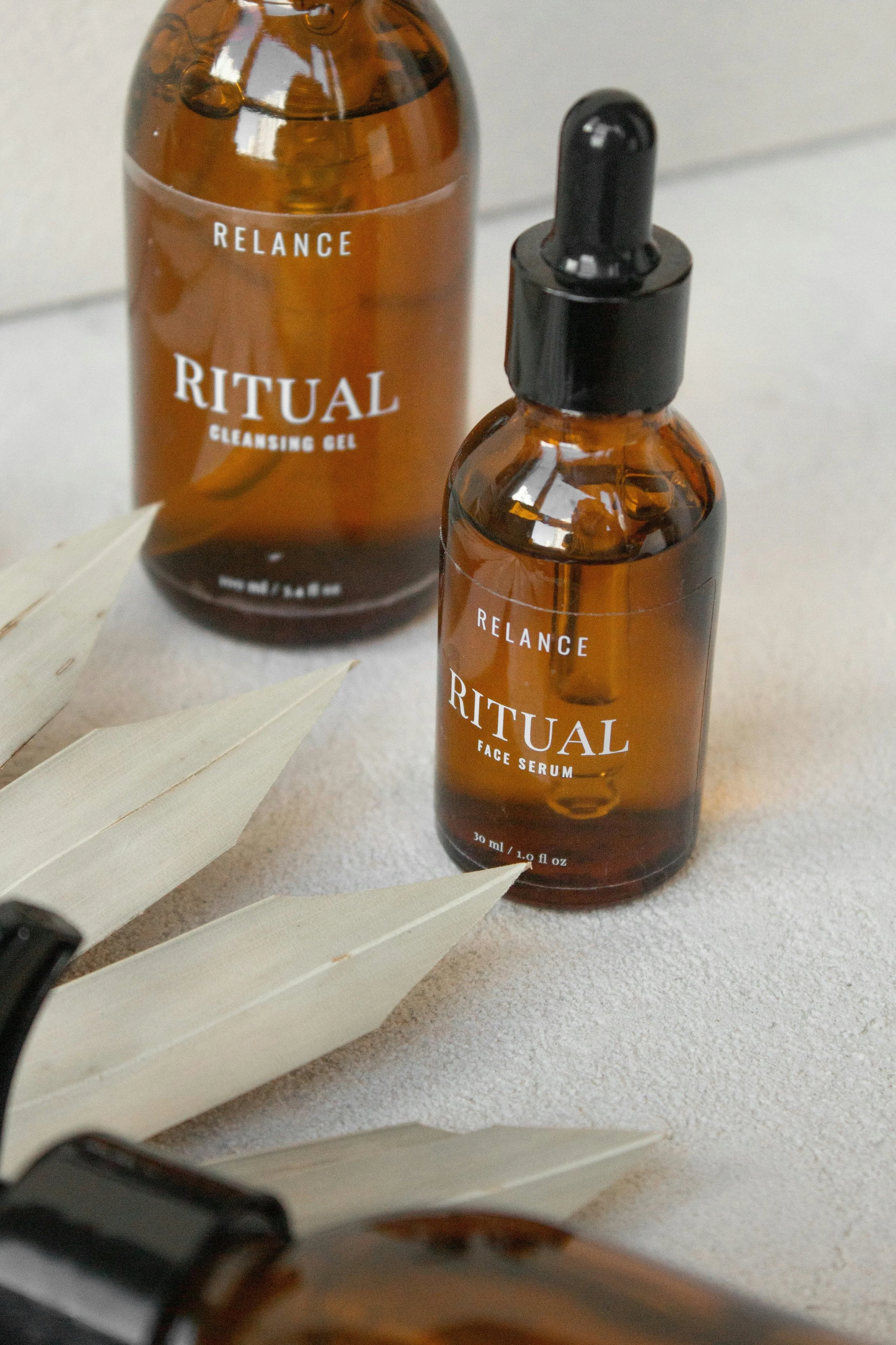 Brown bottles labeled 'Ritual' with one labeled 'Face Serum' and the other 'Cleansing Gel,' set on a light-colored surface next to dried leaves.