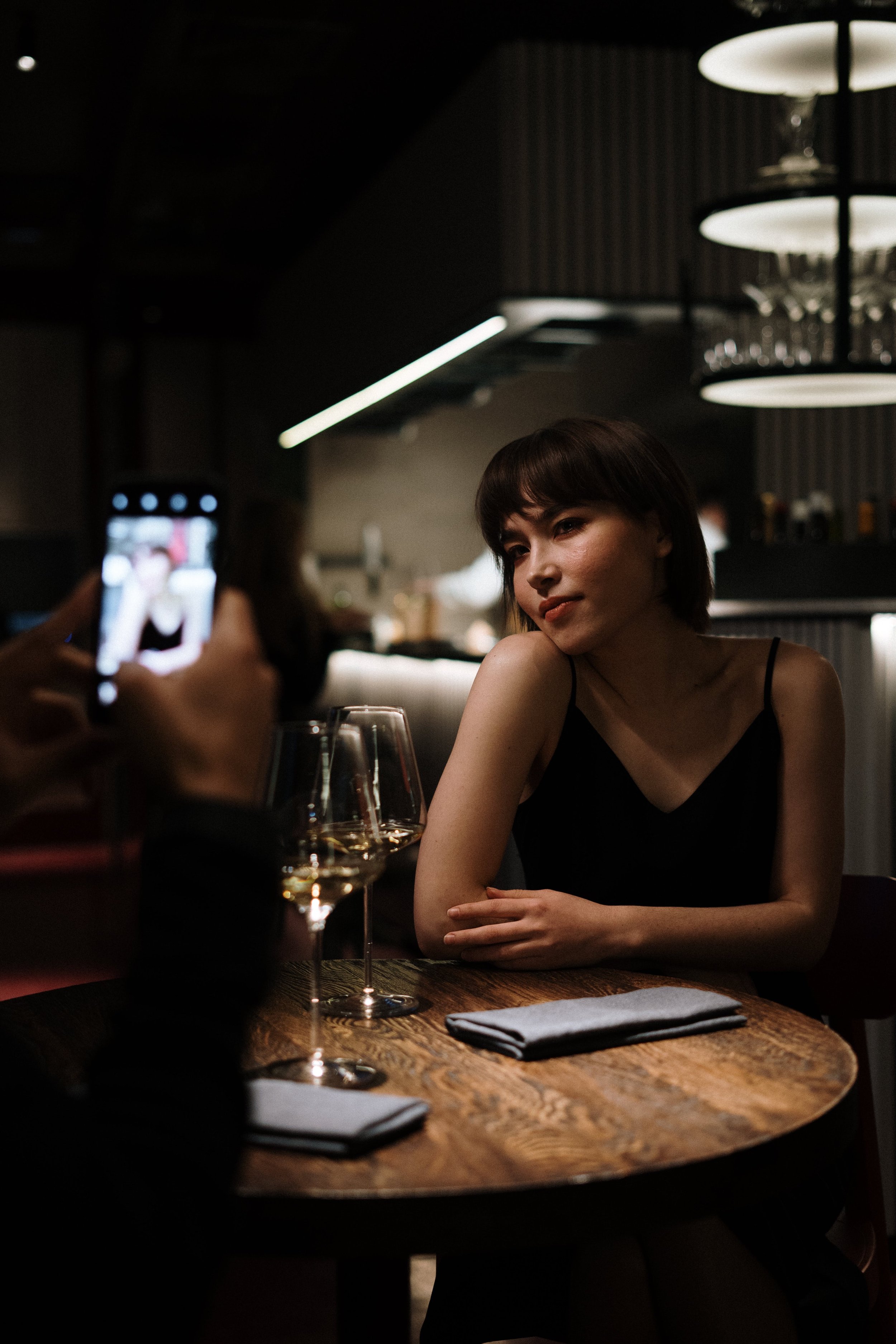 Woman in black dress sitting at a table in a dimly lit restaurant, with two glasses of white wine and a folded napkin in front of her, while someone takes her photo.
