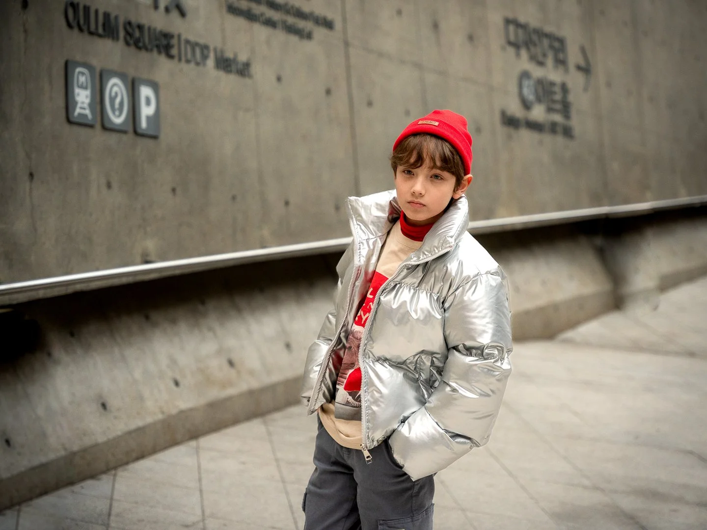 Young boy standing in an indoor parking garage, wearing a silver puffer jacket, red beanie, and casual clothes, with his hands in his pockets, looking slightly to his left.