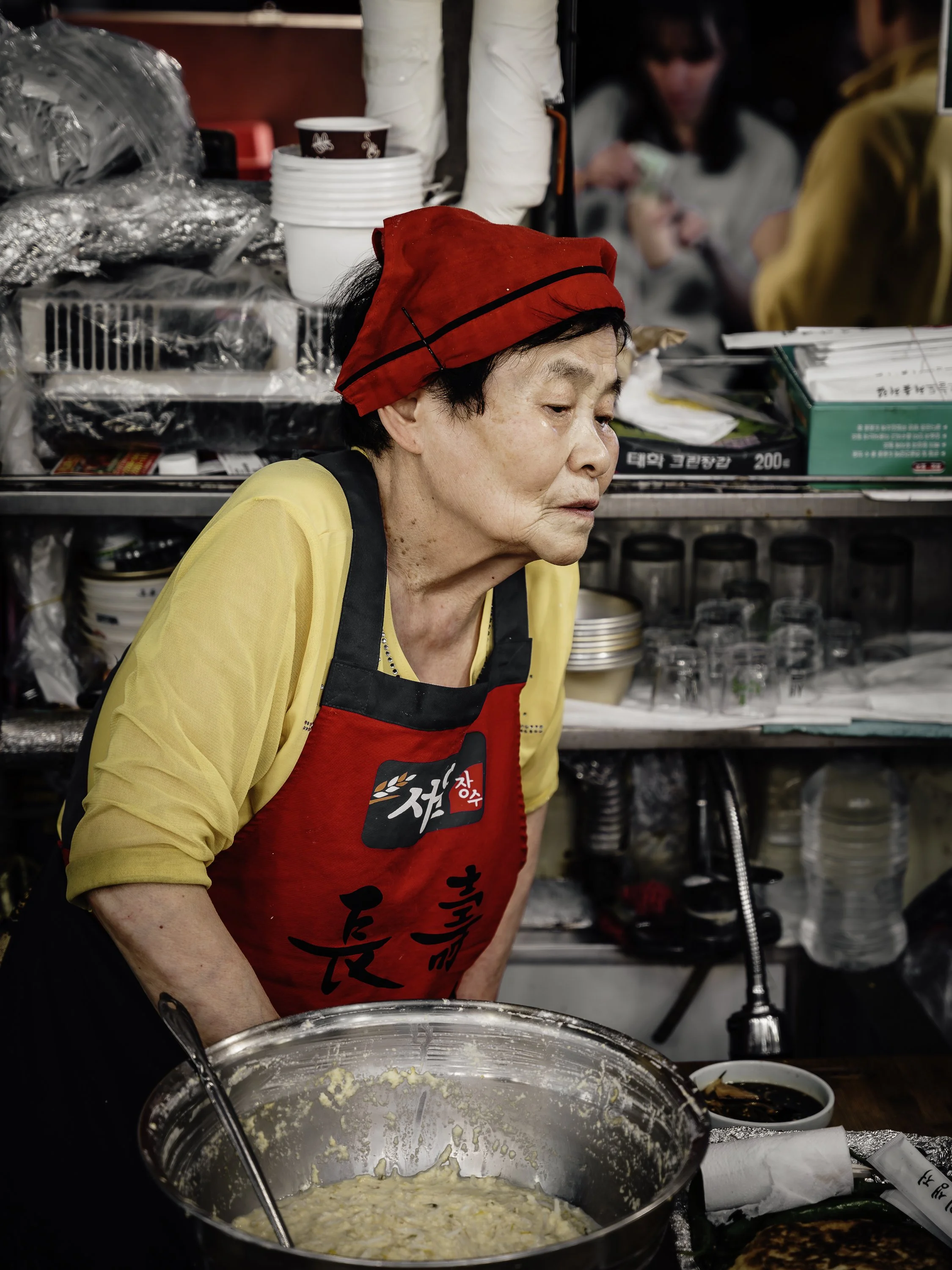 An elderly woman wearing a red headscarf and yellow shirt under a black apron is looking down at a large metal mixing bowl with batter, in a busy kitchen or food stand. There are bowls, plates, and food items around her.