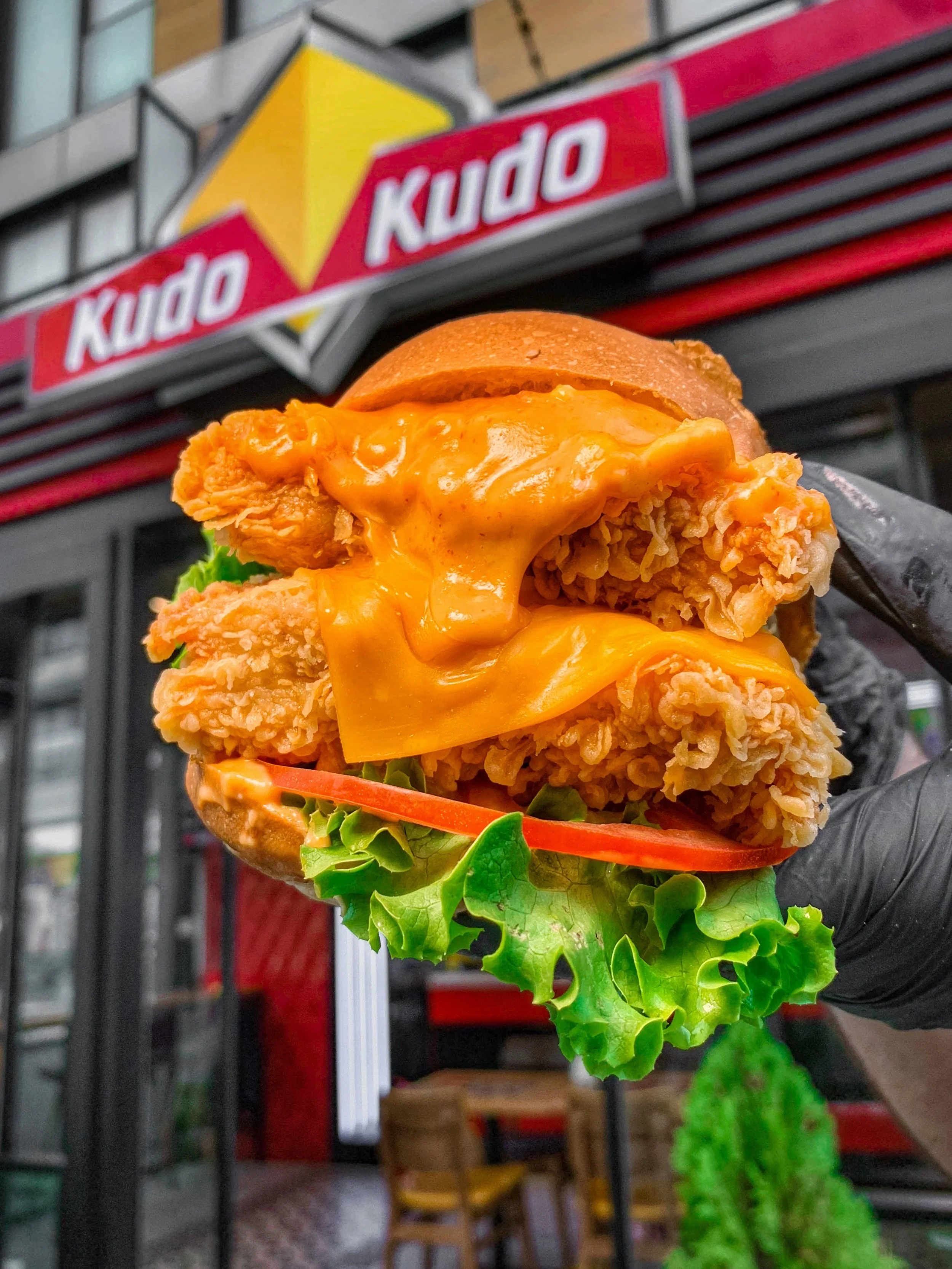 Fried chicken sandwich with cheese, lettuce, and tomato held in front of a Kudo restaurant.