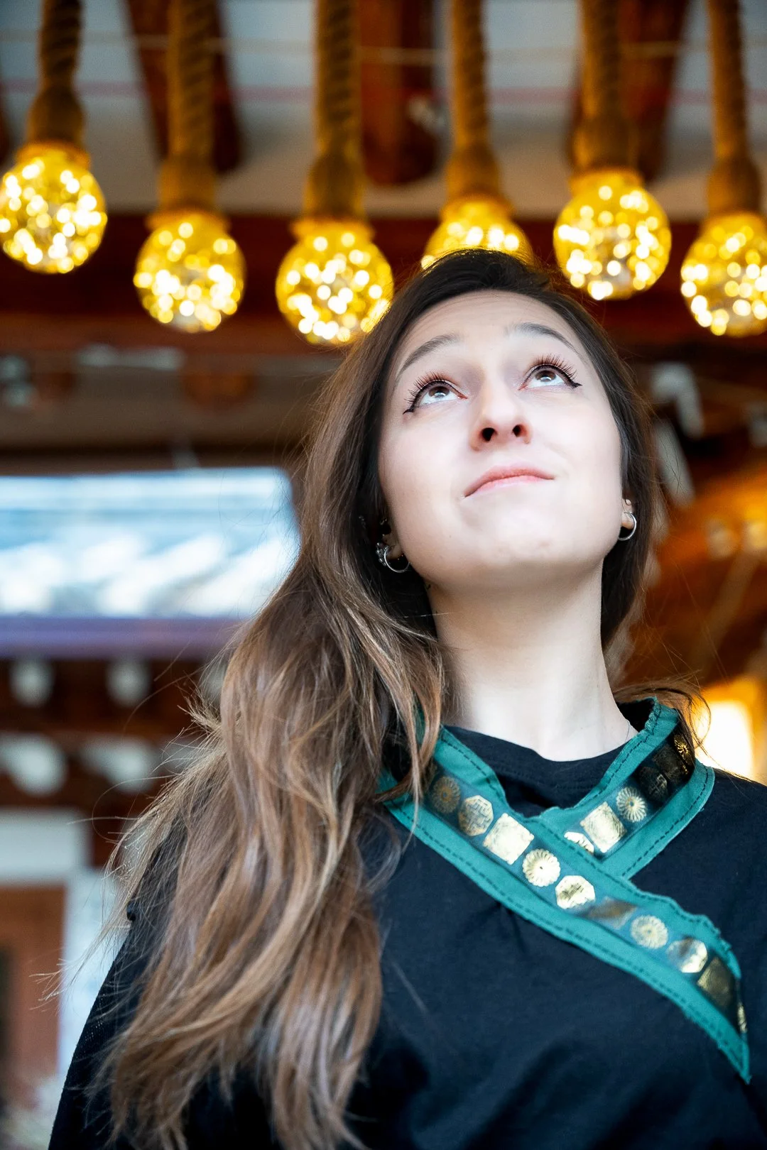A young woman with long wavy hair looking upward. She is wearing earrings and a black outfit with green and gold embellishments around the collar. The background features warm yellow hanging lights and a blurred interior setting.
