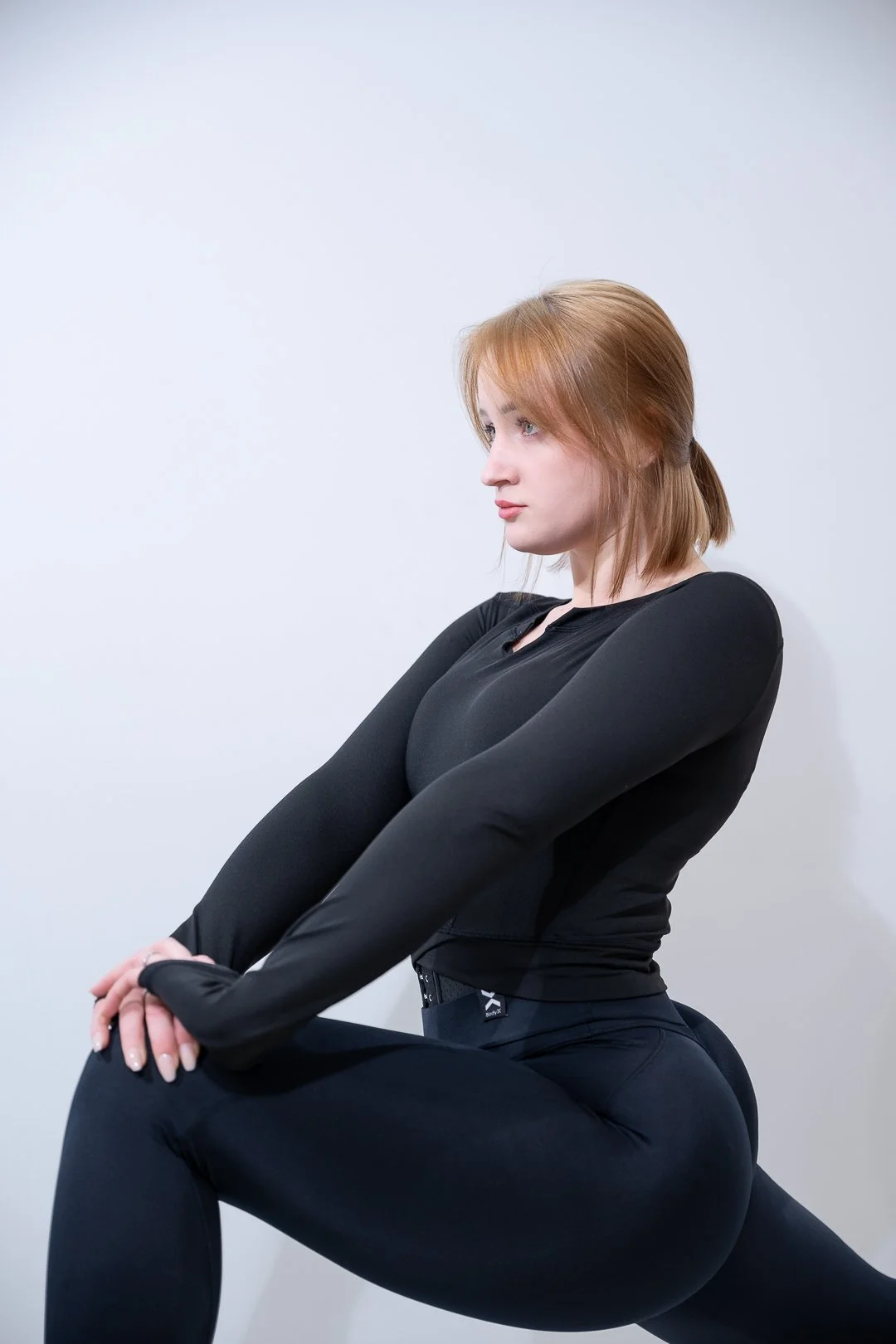 A woman with shoulder-length red hair in black athletic wear is stretching against a plain white wall.