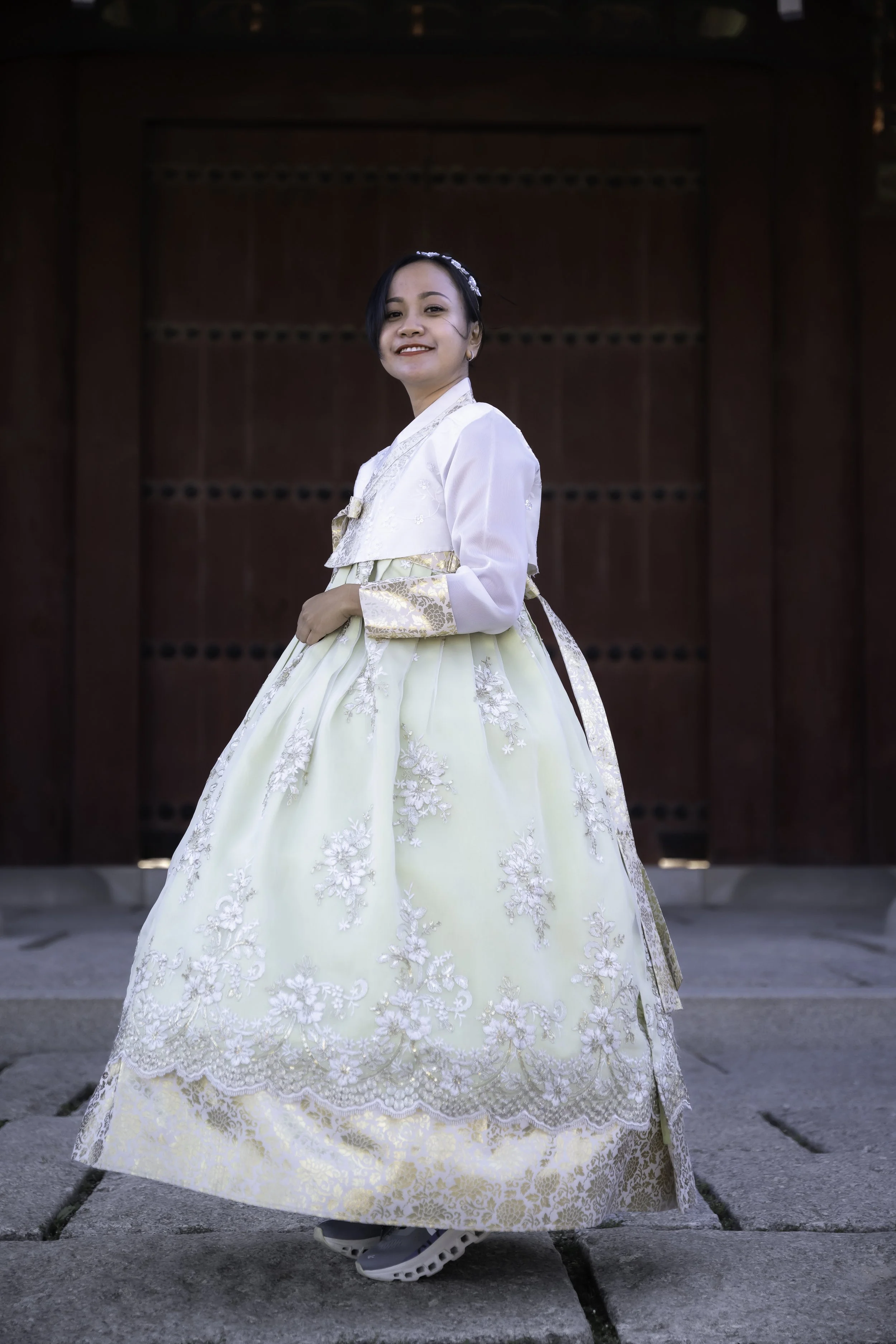 A woman wearing a traditional white and cream dress stands in front of a red wooden door, smiling at the camera.