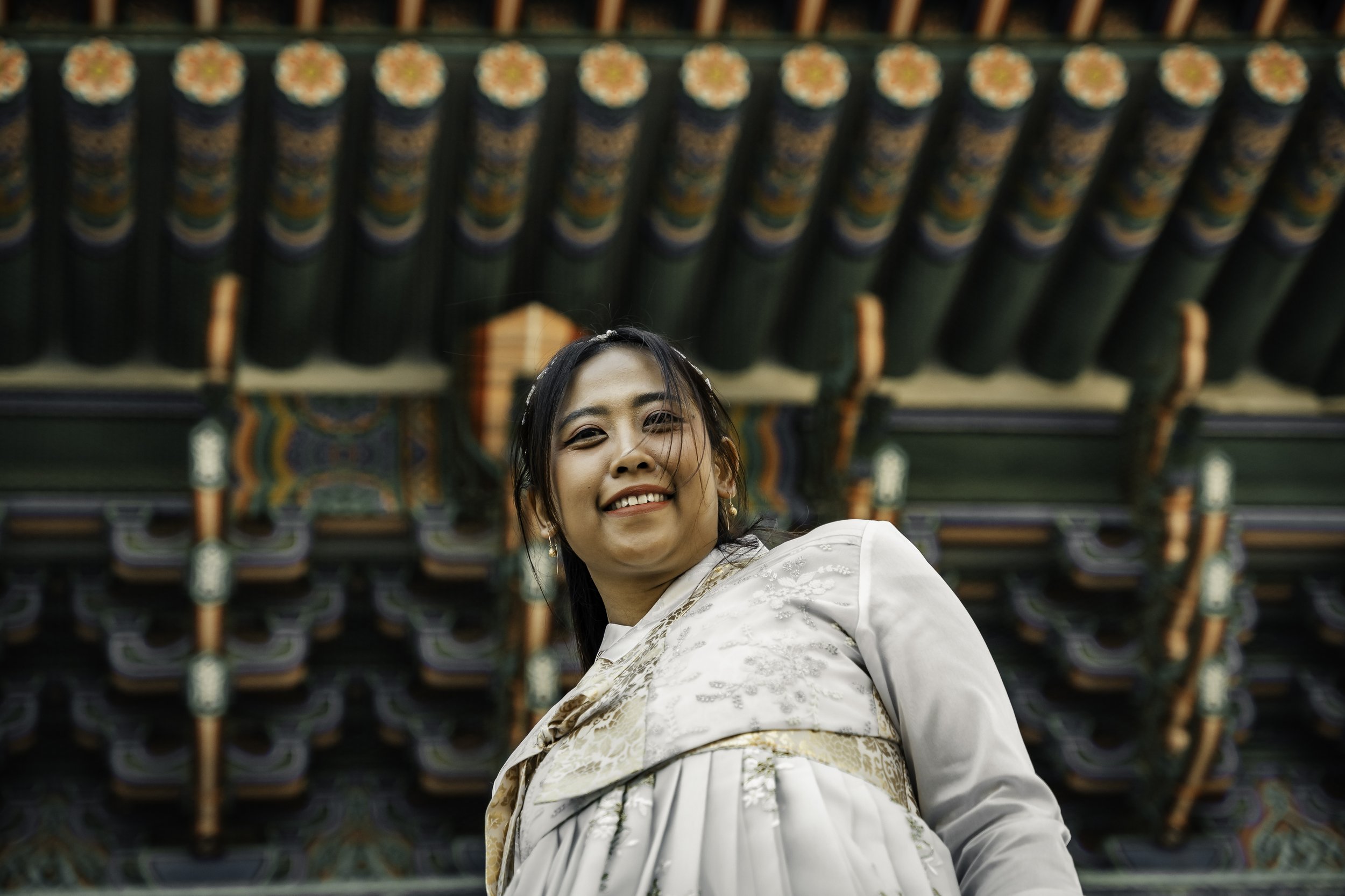 A smiling woman in traditional Korean hanbok standing in front of a colorful, ornate building roof.