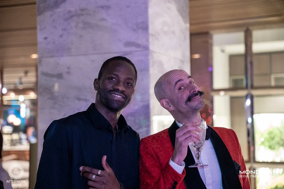 Two men at a social event, one wearing a black shirt and the other in a red suit jacket with a glass in hand, posing and smiling.